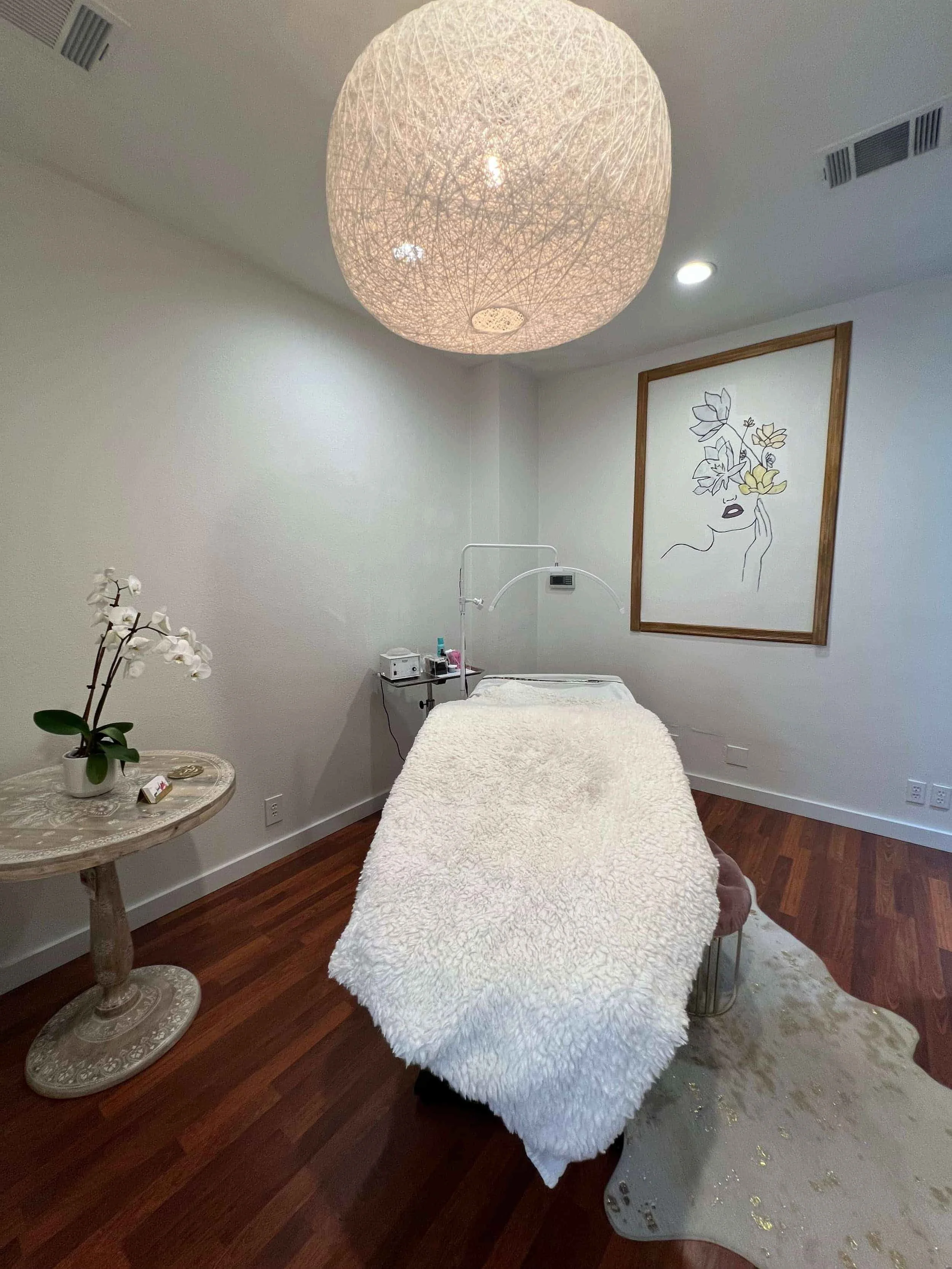 A cozy spa room with a massage bed covered in a fluffy white blanket, a round side table with a white orchid plant, and wall art of a woman's face with floral accents. There is a large spherical ceiling light and a framed art piece on the wall.