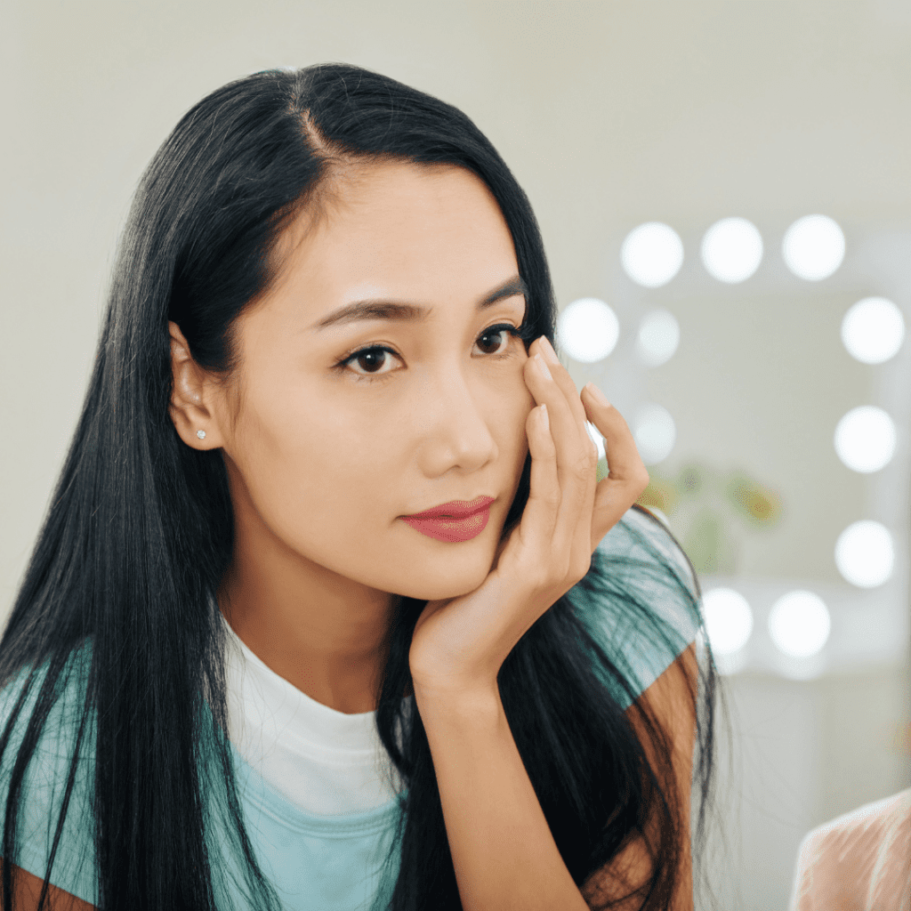 Close-up of a young woman with long black hair, wearing a white top with teal sleeves, touching her face gently, with a background of bright vanity lights.