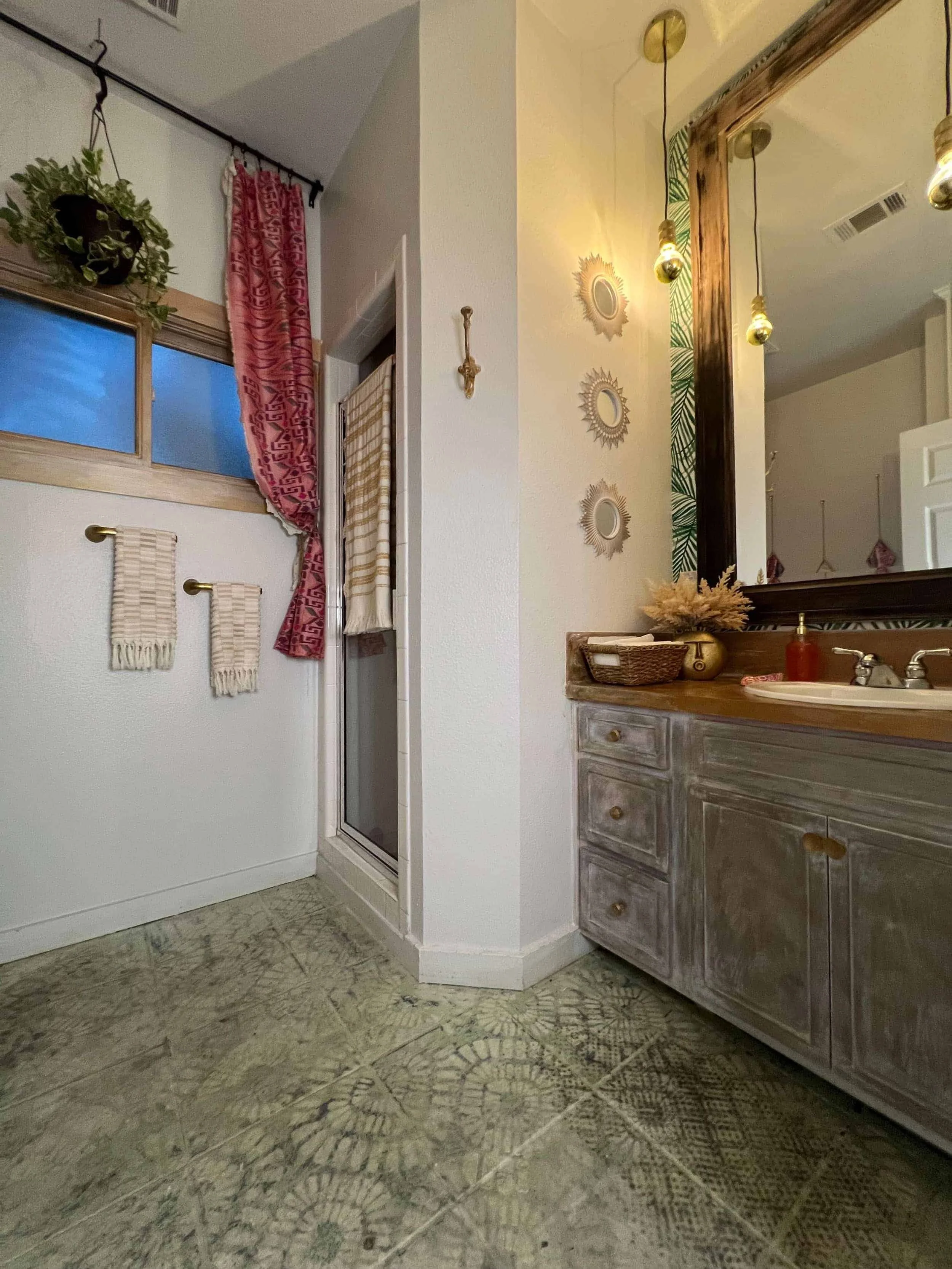 Bathroom with a tiled floor, a vanity with a large mirror, hanging pendant lights, decorative mirrors, towels on hooks, a window with a pink curtain, and plants.