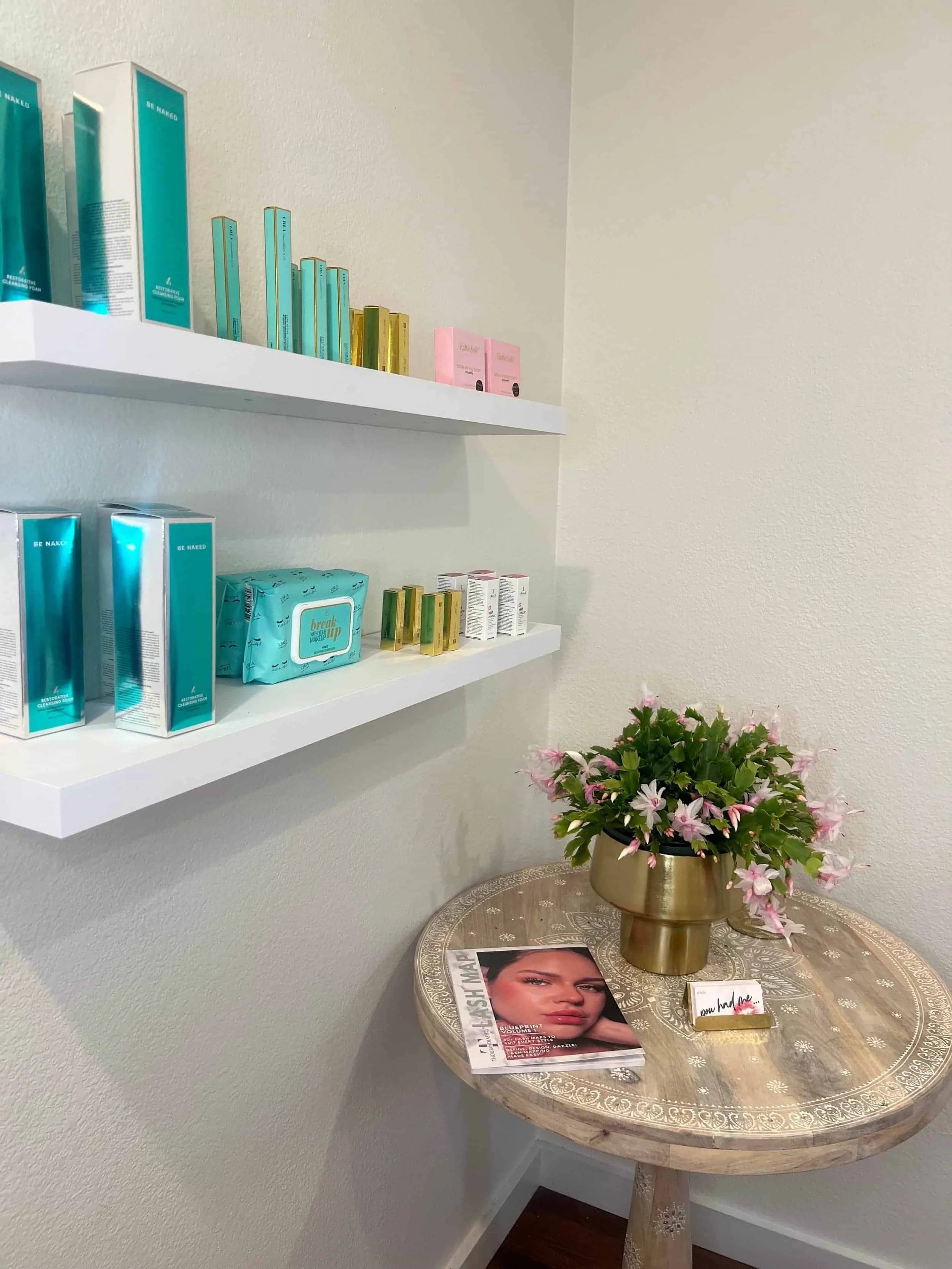 A round wooden table with a vase of pink flowers, a fashion magazine, and a small box. Above the table, white shelves hold skincare products and books.
