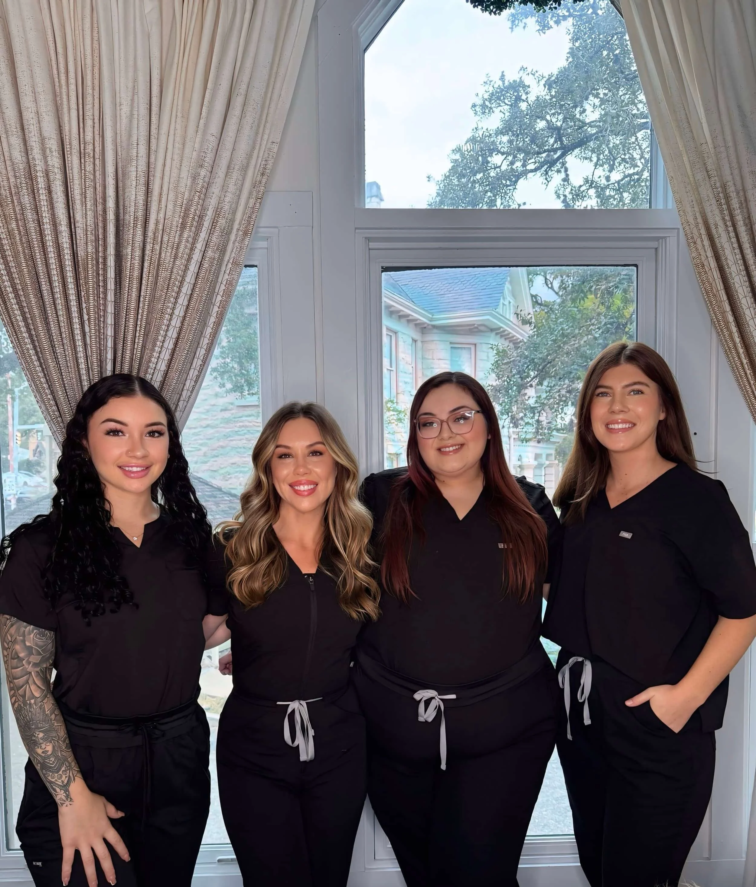 Four women standing side by side indoors in front of a large window with curtains, wearing matching black uniforms with gray drawstrings, smiling at the camera.