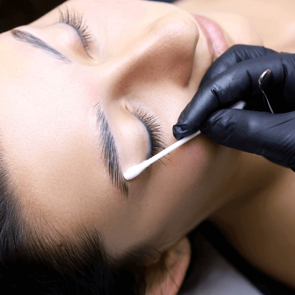 A person receiving eyelash cleaning or eyelash extension treatment, lying down with eyes closed, while a technician in black gloves uses a cotton swab near the eye.