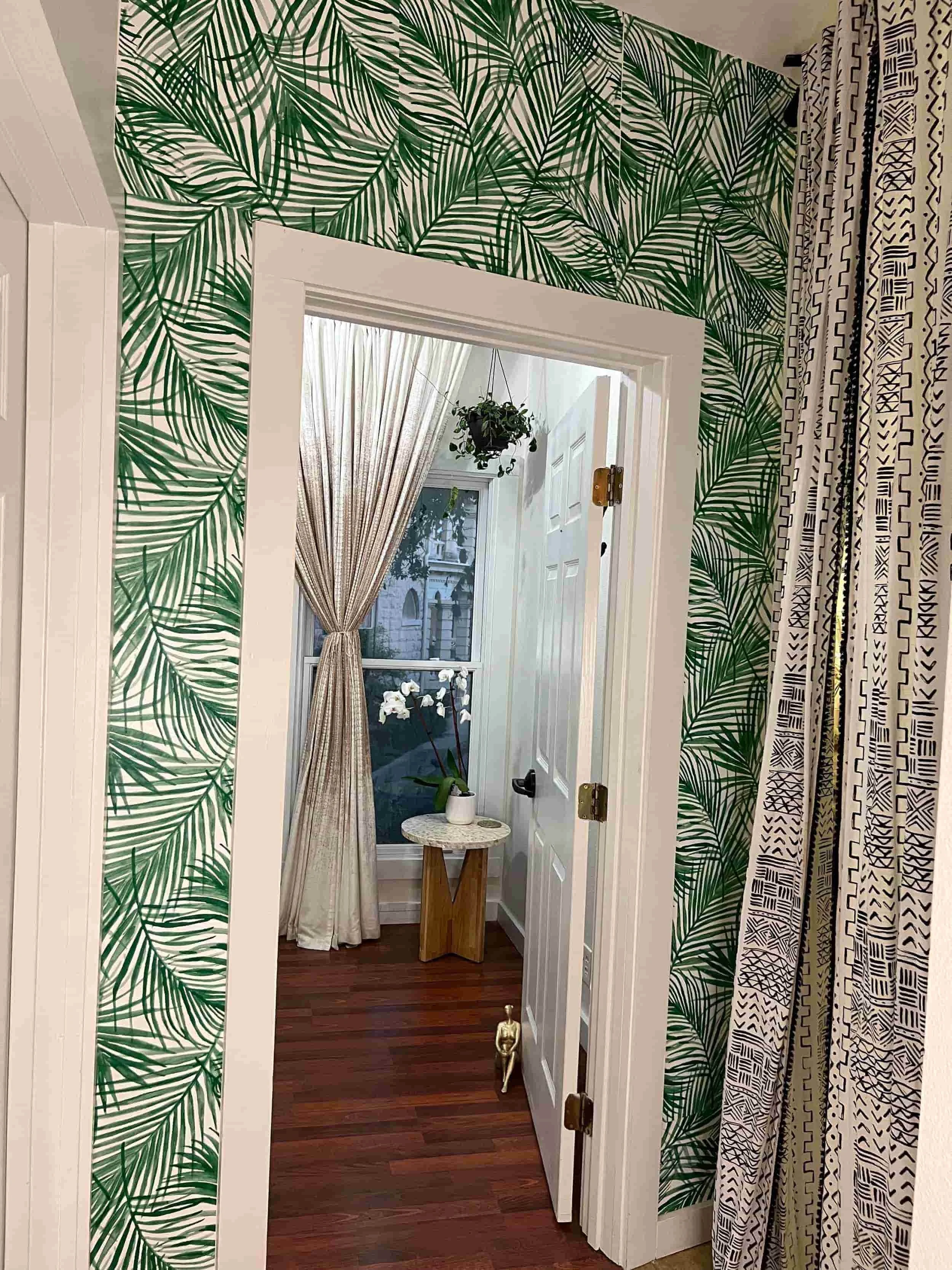Interior view of a room with tropical leaf wallpaper, white trim, a window with curtains, a small side table with a potted plant, and hardwood floors.
