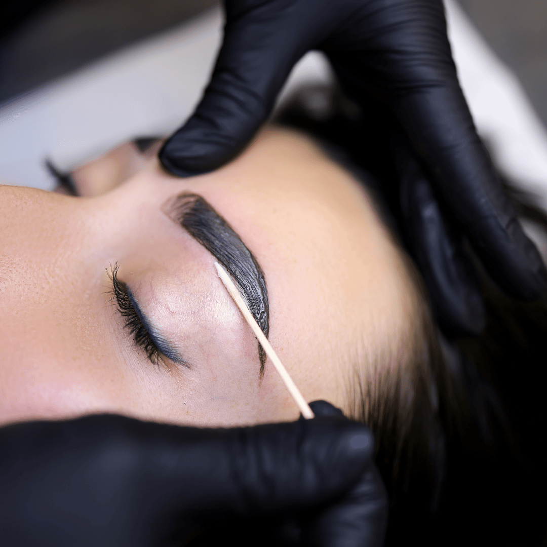 A person receiving eyelash extension or eyelash tinting treatment, with a beautician wearing black gloves applying product to the person's closed eyelid using a small stick.