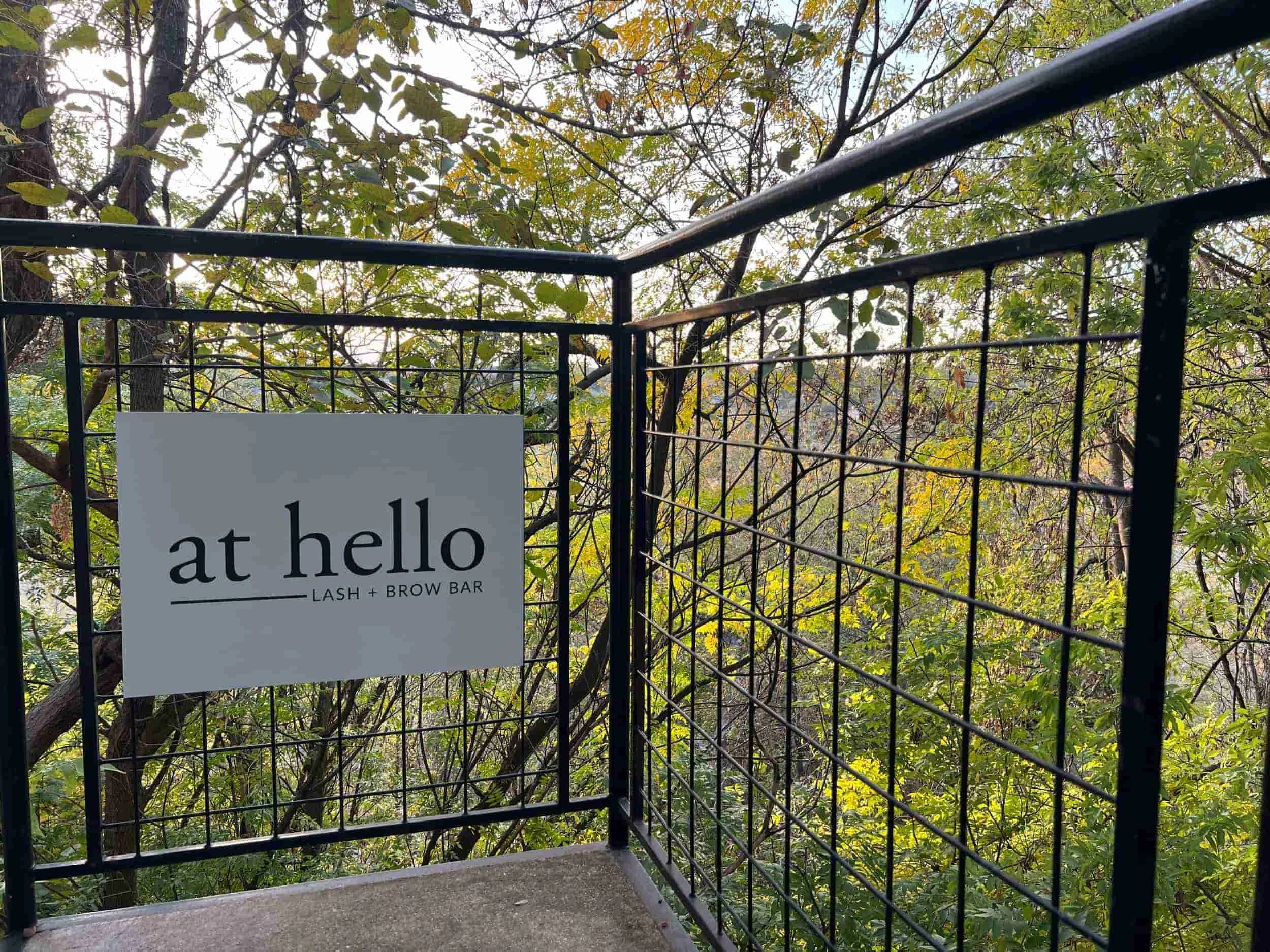 Outdoor balcony with a black metal railing and a sign that reads 'at hello Lash + Brow Bar', overlooking a green wooded area with trees and leaves.