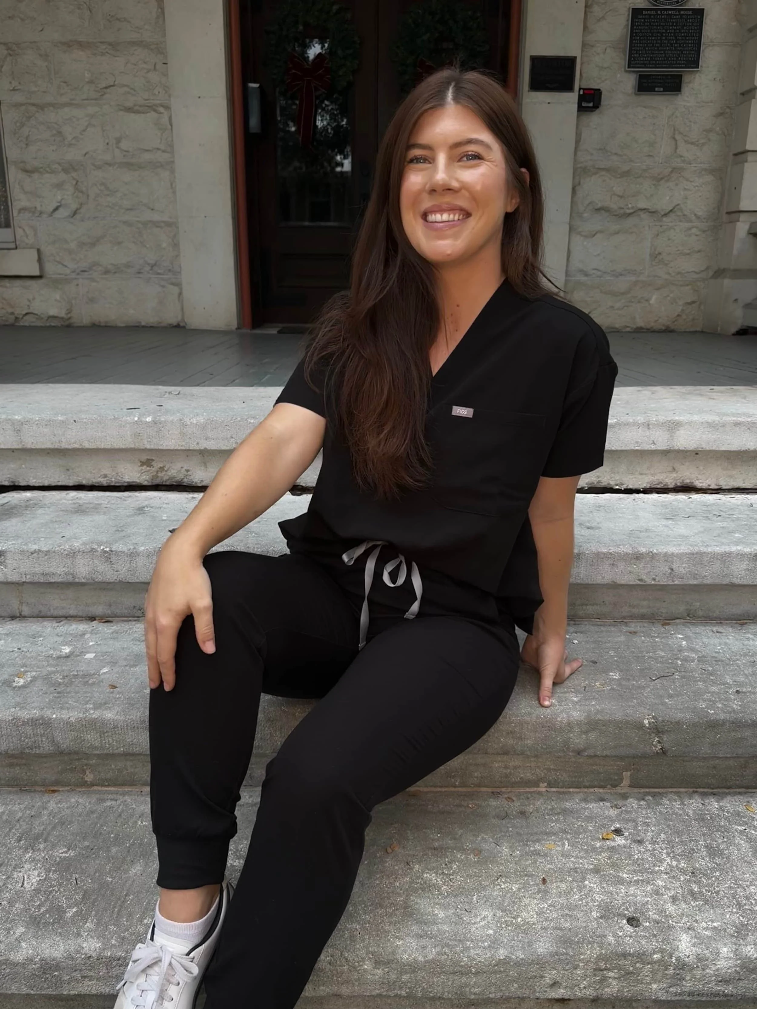 Woman with long dark hair sitting on stone steps outside a building, smiling, wearing black scrubs, white sneakers, and a white drawstring at the waist.