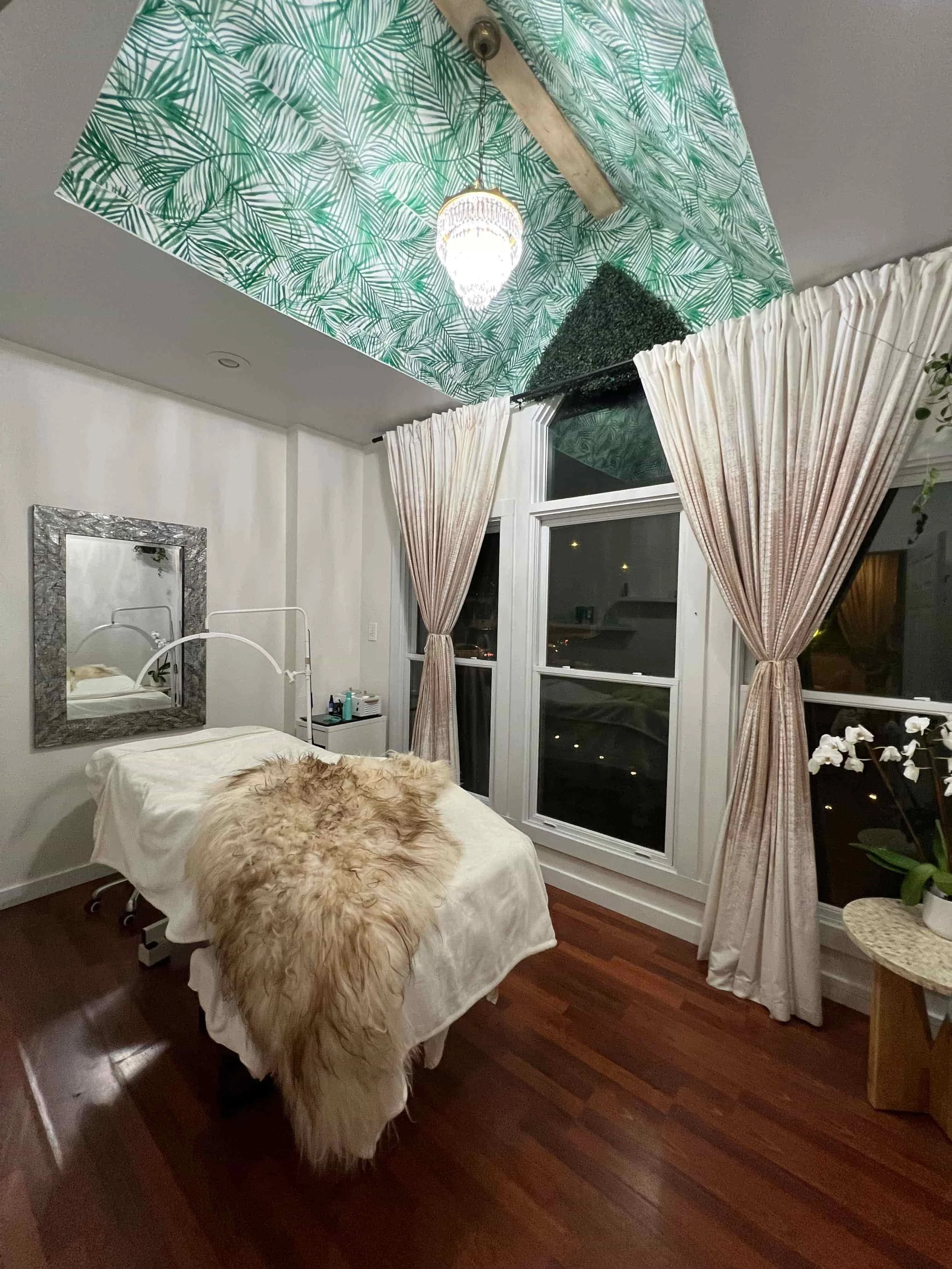 A medical or spa treatment room with a massage table covered in a white sheet and a furry beige blanket. There is a large window with beige curtains, a mirror on the wall, and a small round table with a potted orchid plant. The ceiling is decorated w