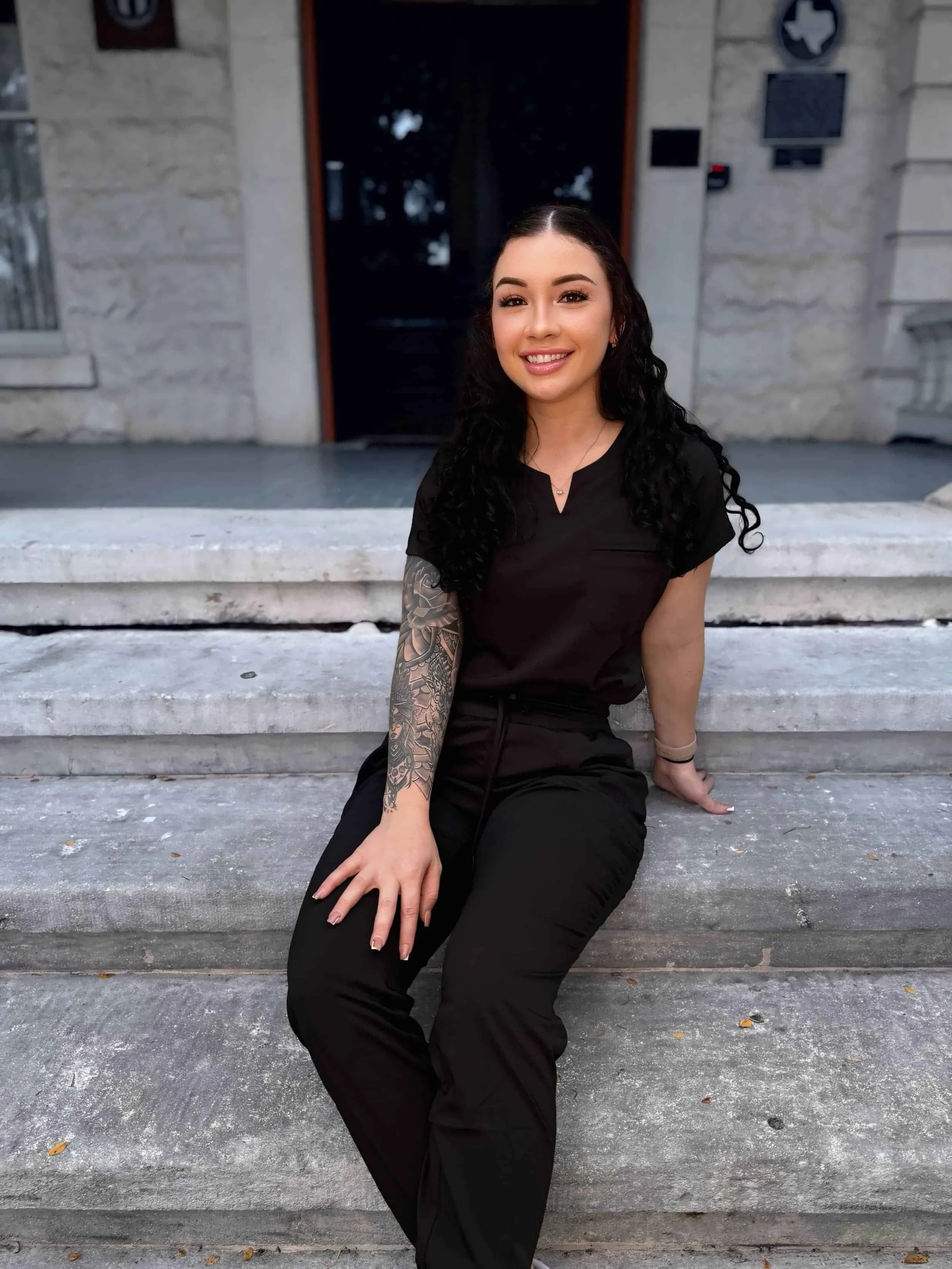 A young woman with long curly black hair sitting on stone steps outside a building, smiling, wearing a black scrubs uniform, with a tattoo sleeve on her left arm.