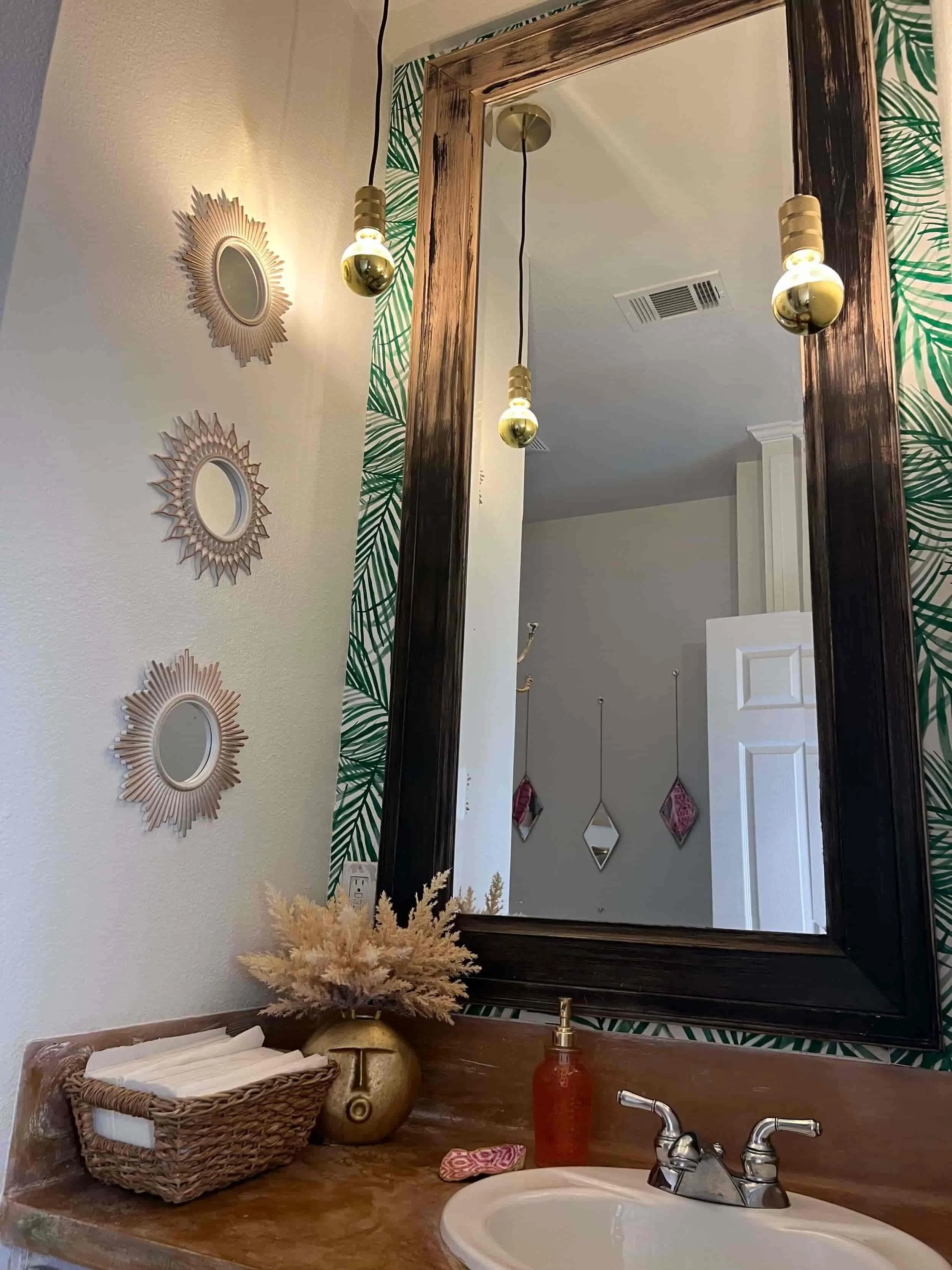 Bathroom vanity with a large mirror, hanging pendant lights, decorative wall mirrors, a basket with towels, a vase with dried flowers, soap, and a sink.