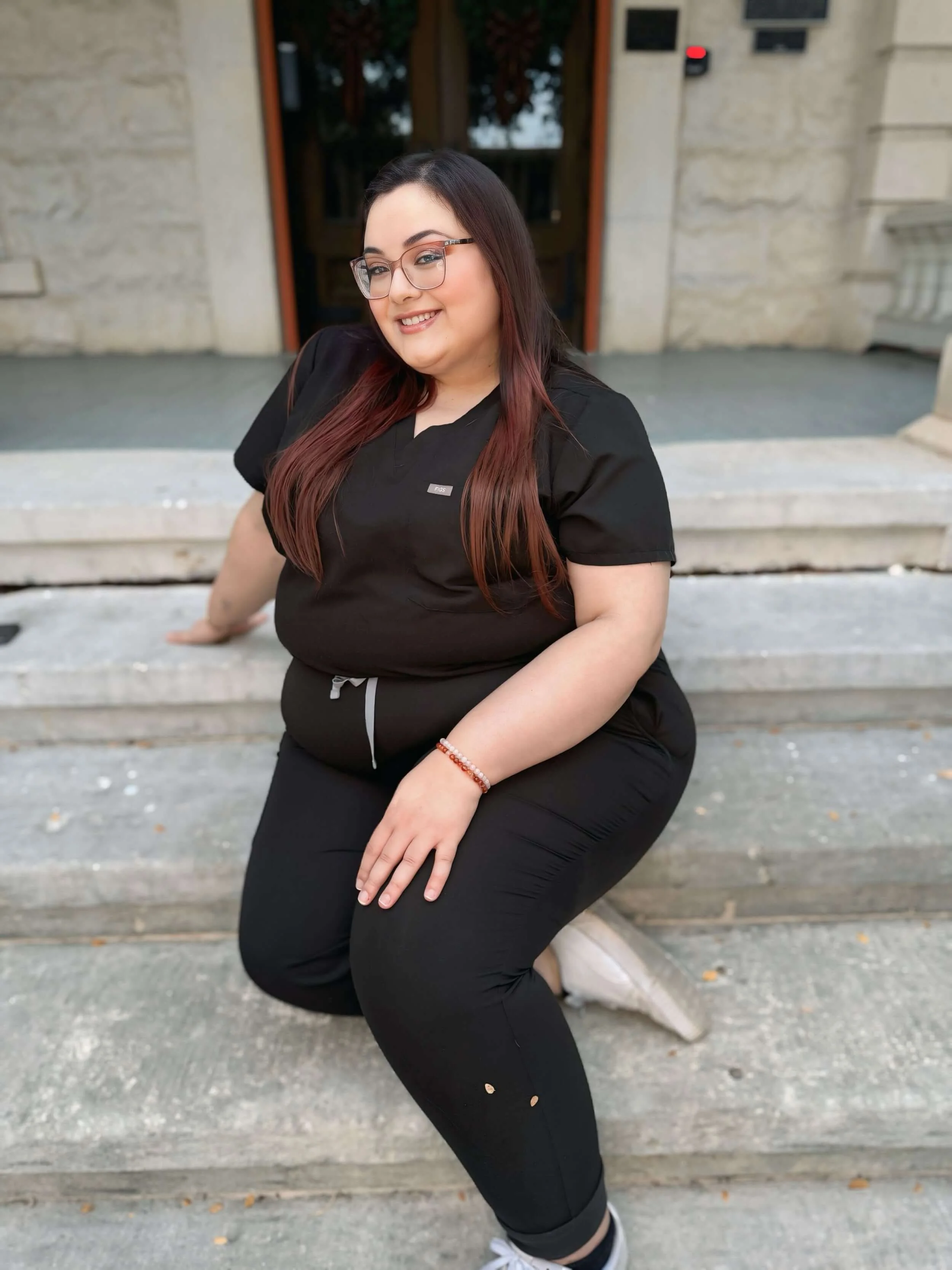 A woman kneeling on a stone step outside a building, smiling and wearing black scrubs with glasses and a beaded bracelet.