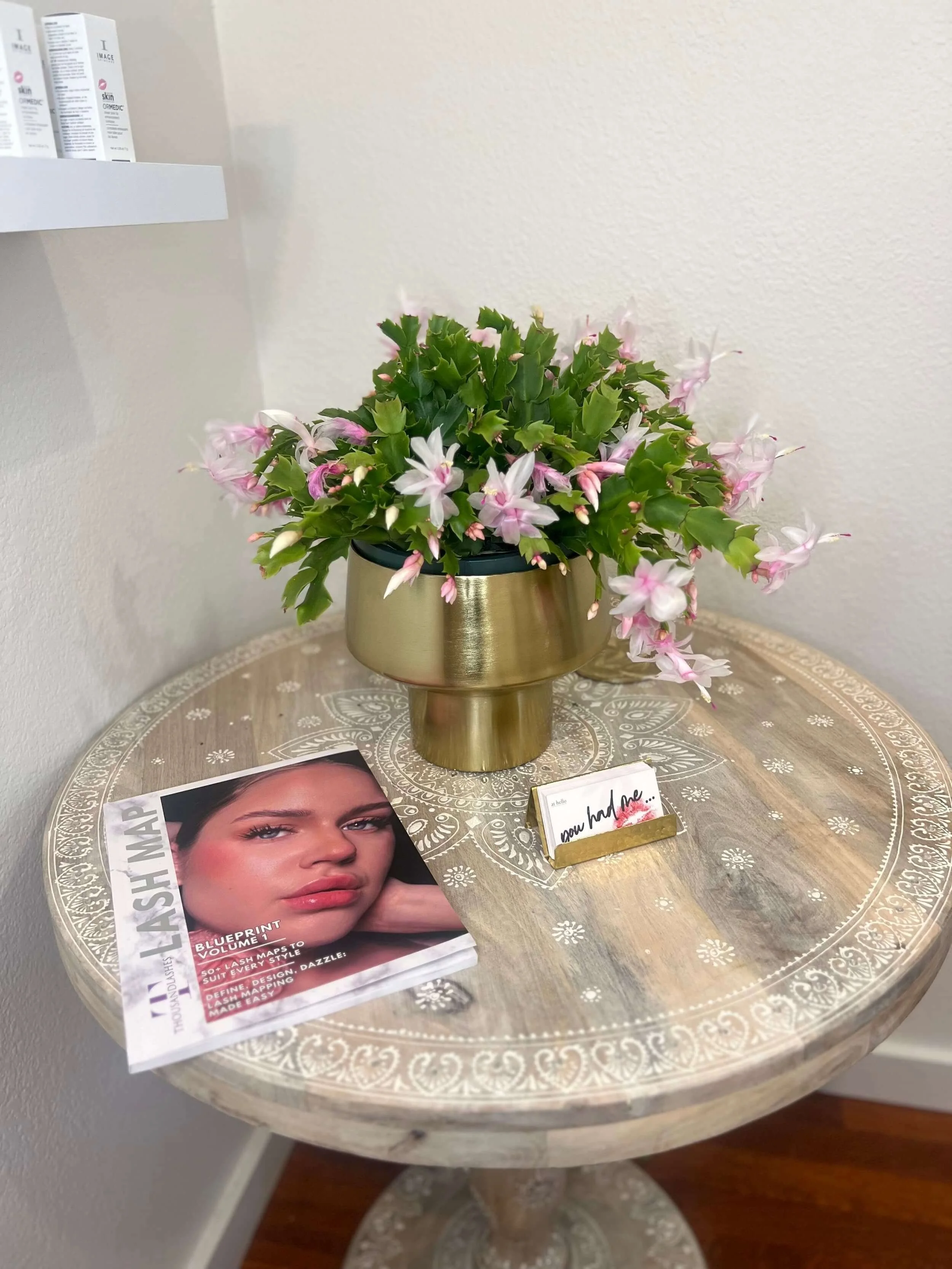 Decorative round wooden table with white painted floral design, holding a pink and white flowering plant in a gold-colored vase, a beauty magazine featuring a woman with makeup on the cover, and a small white and gold display card with black cursive 