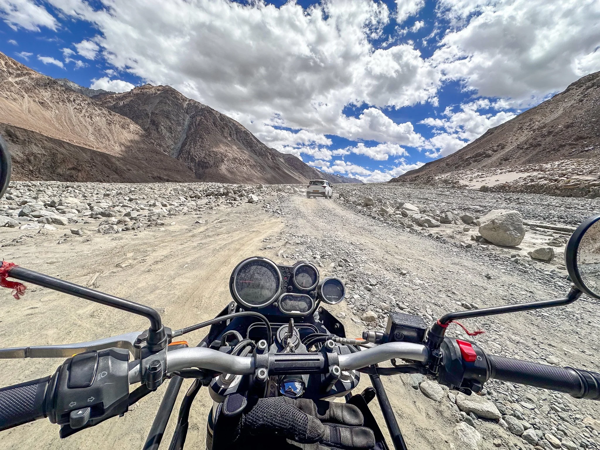 View from a motorcycle rider's perspective on a rocky dirt road in a mountainous area with a car ahead and partly cloudy sky above.