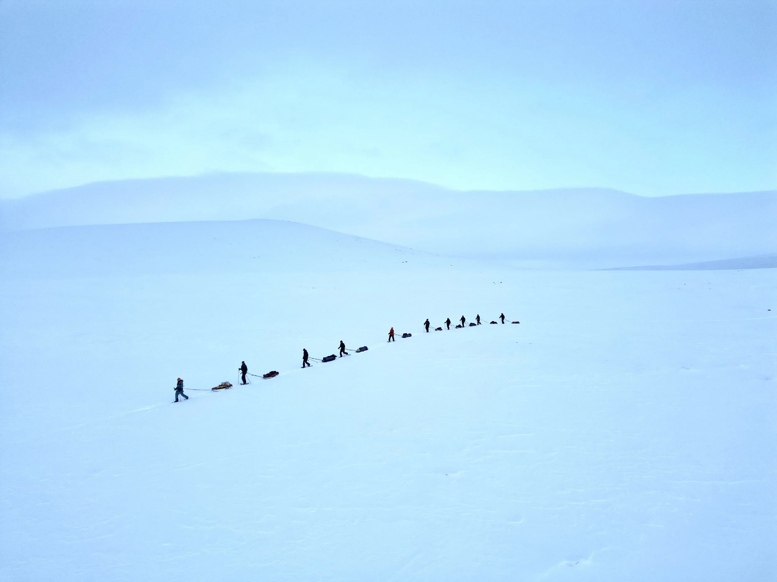 A group of people trekking across a vast, snow-covered landscape with mountain ranges in the distance under a pale blue sky.