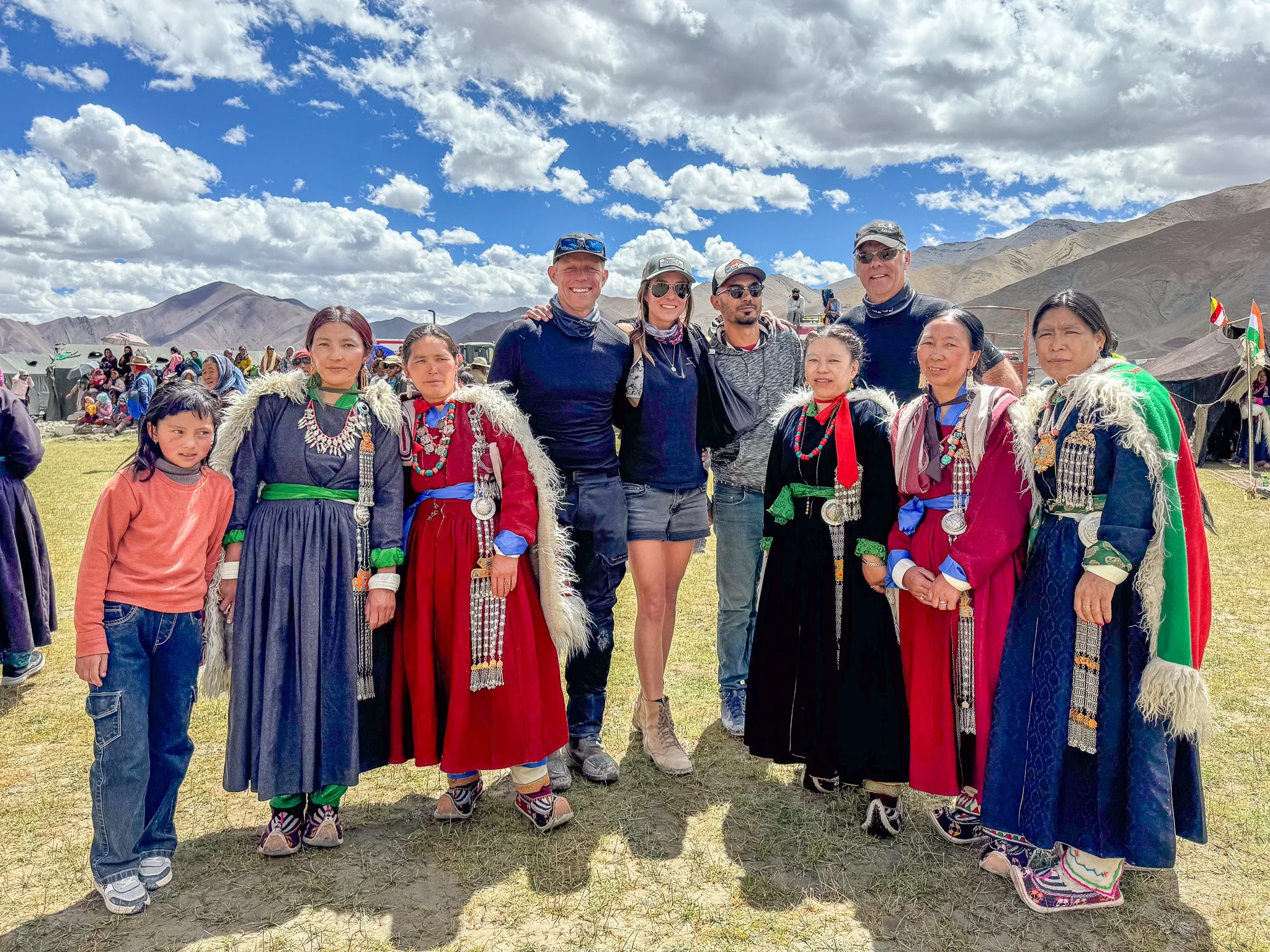 Group of people standing outdoors, some wearing traditional clothing with colorful jewelry and embroidery, in a mountainous area with partly cloudy blue sky.