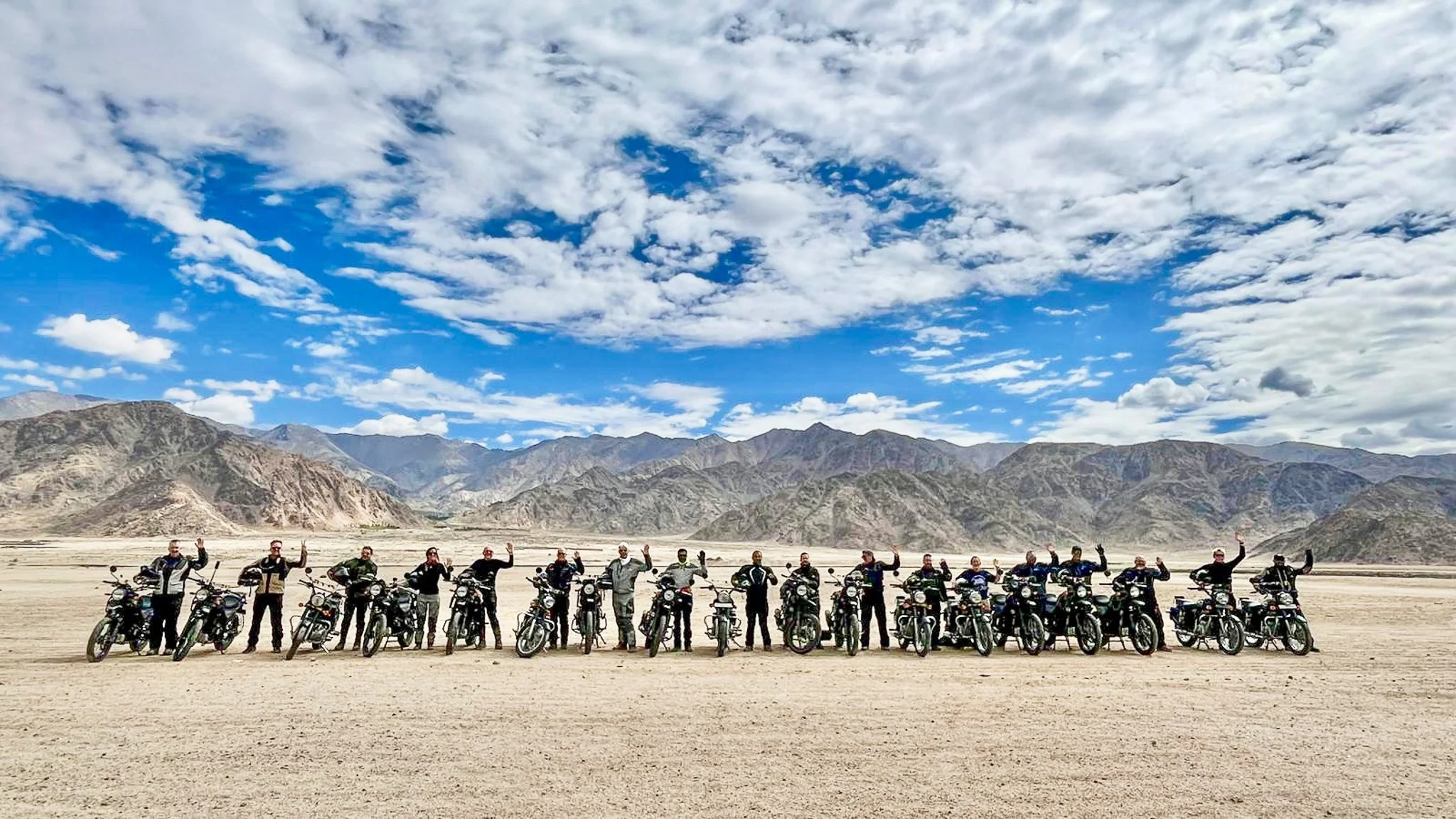 A group of people standing beside their motorcycles in a desert landscape with mountains in the background and a partly cloudy sky.