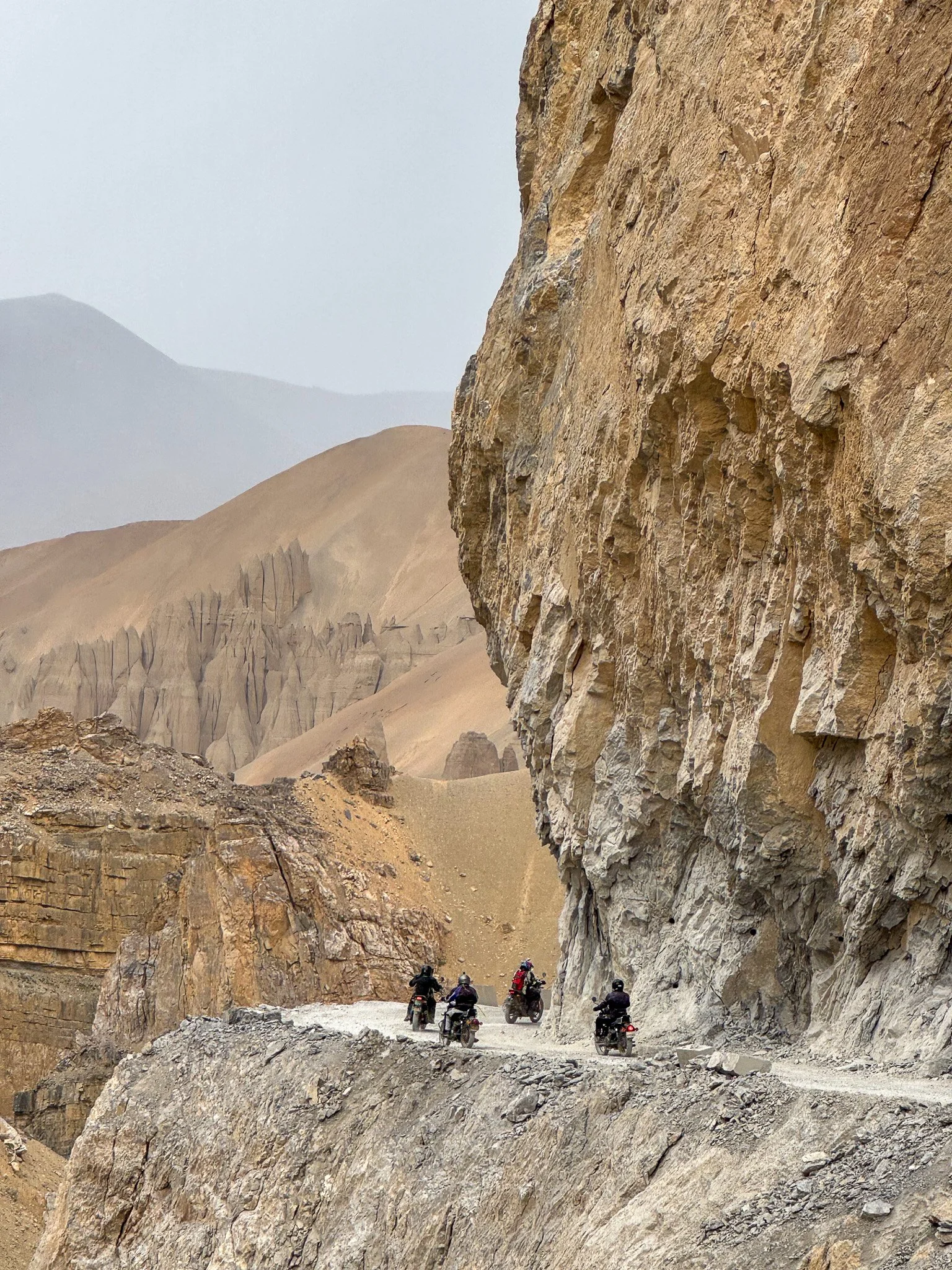 Six motorcyclists riding through a rocky mountain pass with tall cliffs on both sides and barren desert terrain in the background.