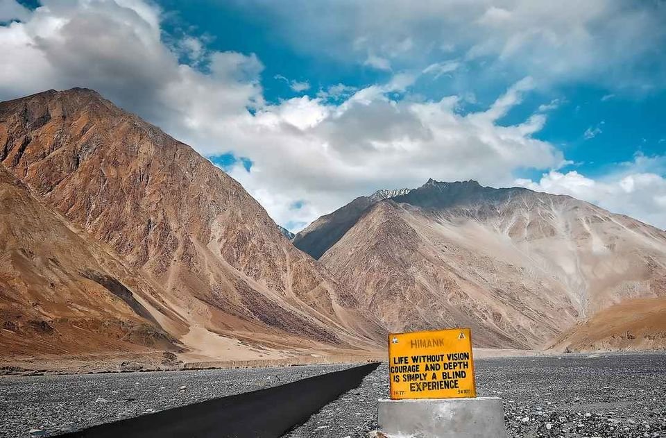 A mountainous landscape with a partly cloudy sky and a sign on the road that reads: 'HIMANK. Life without vision, courage and depth is simply a blind experience'.