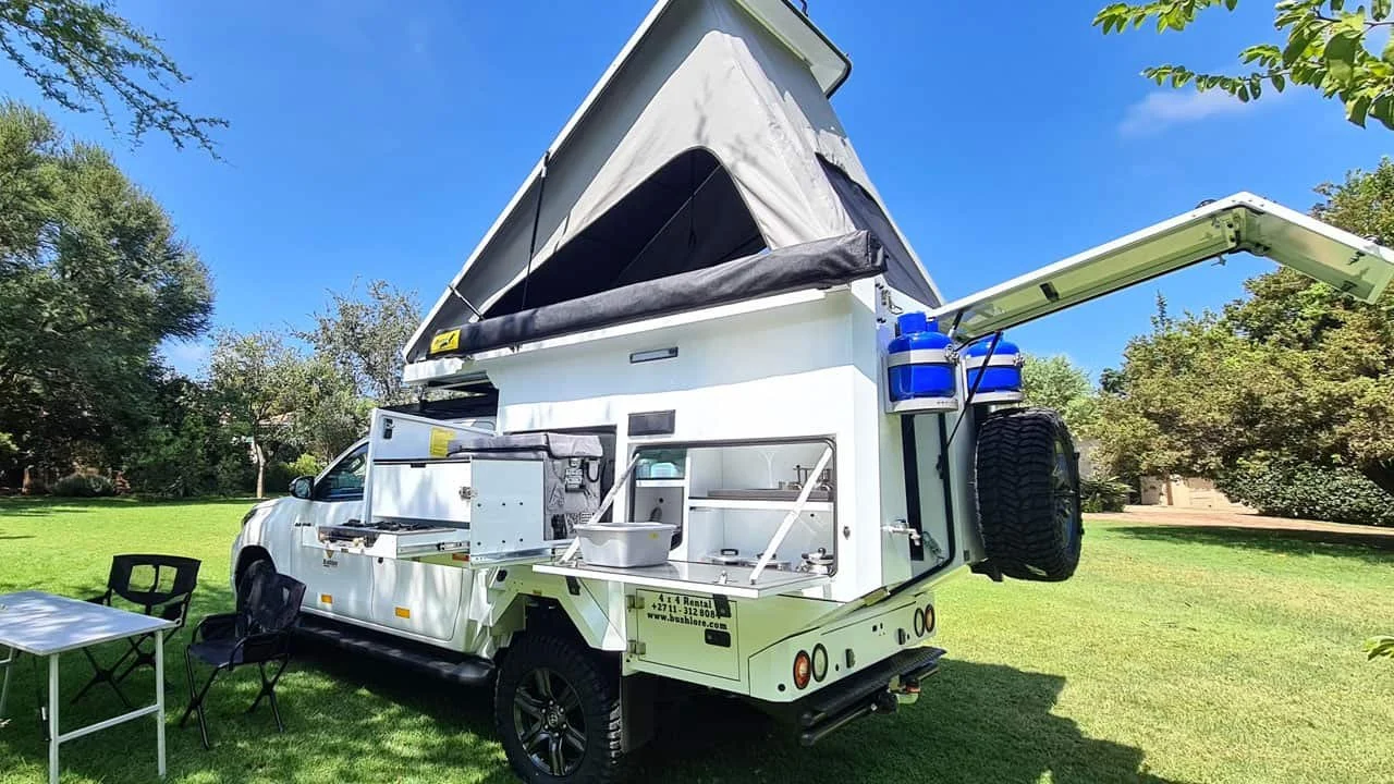 A white overland camper truck with a pop-up roof, outdoor kitchen setup, and storage compartments is parked on a grassy area with trees under a blue sky.