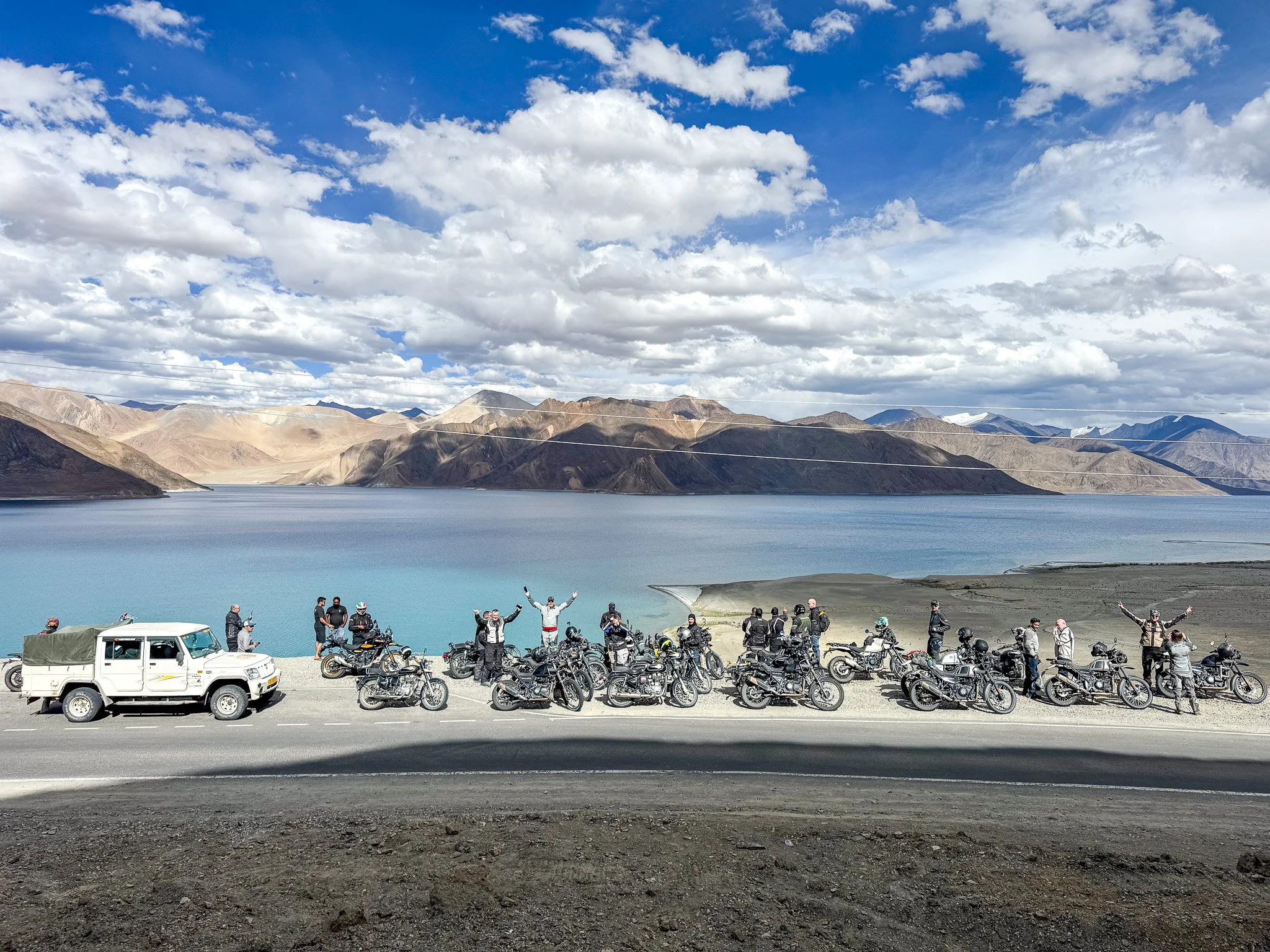 Group of motorcyclists and vehicles parked near a large lake surrounded by mountains under a partly cloudy sky
