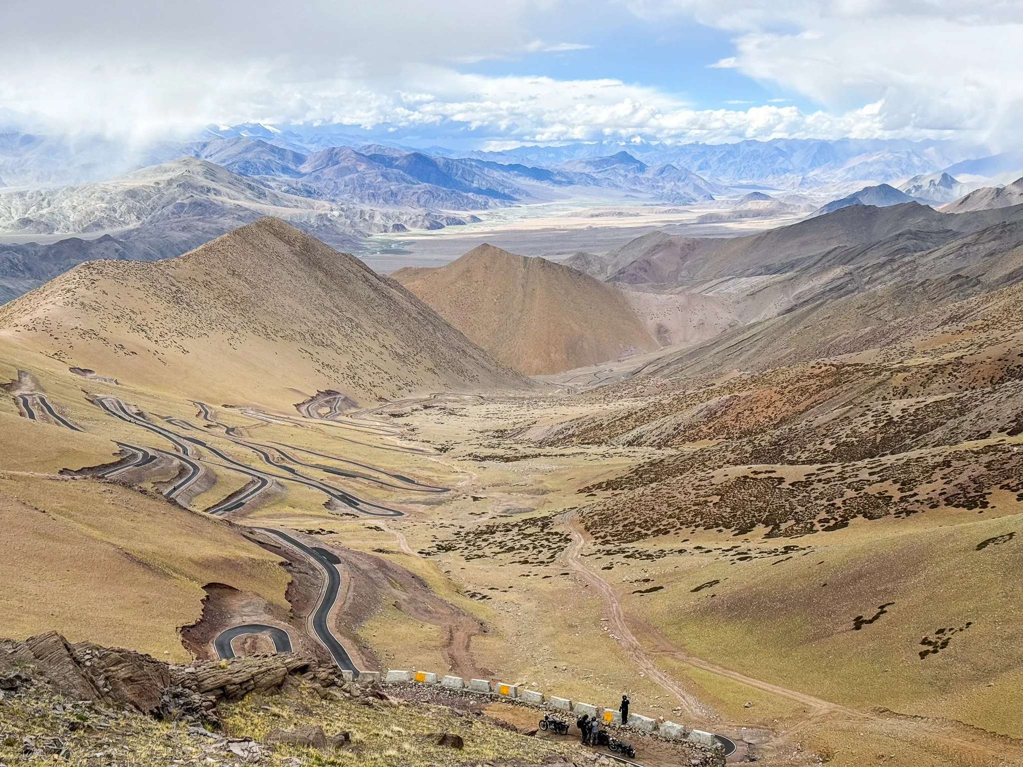 Winding mountain road with a few motorcycles and people at the bottom, surrounded by arid hills and distant snow-capped mountains under cloudy sky.