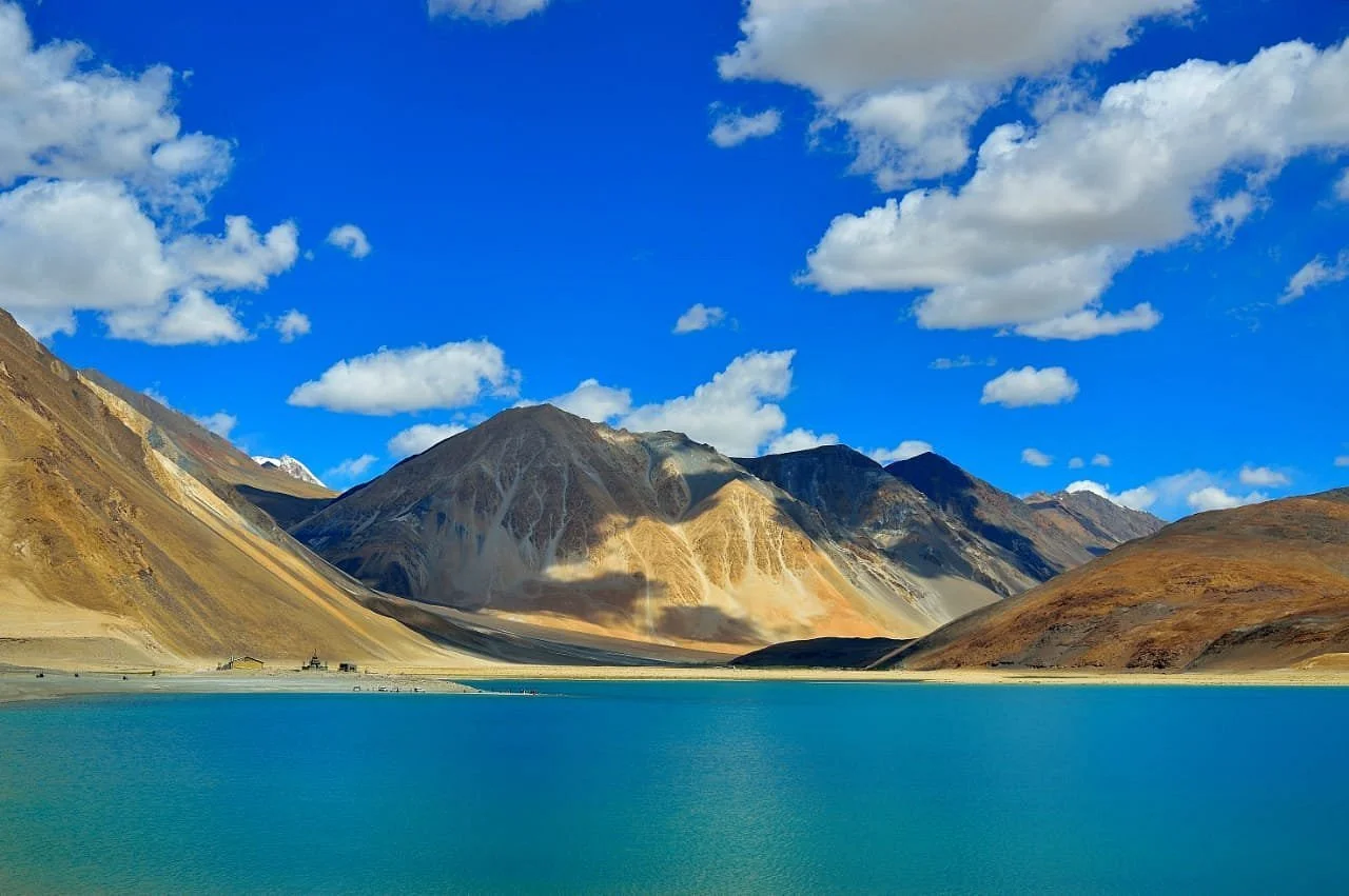 Scenic view of a turquoise lake surrounded by mountains with brown and gray slopes under a blue sky with scattered white clouds.