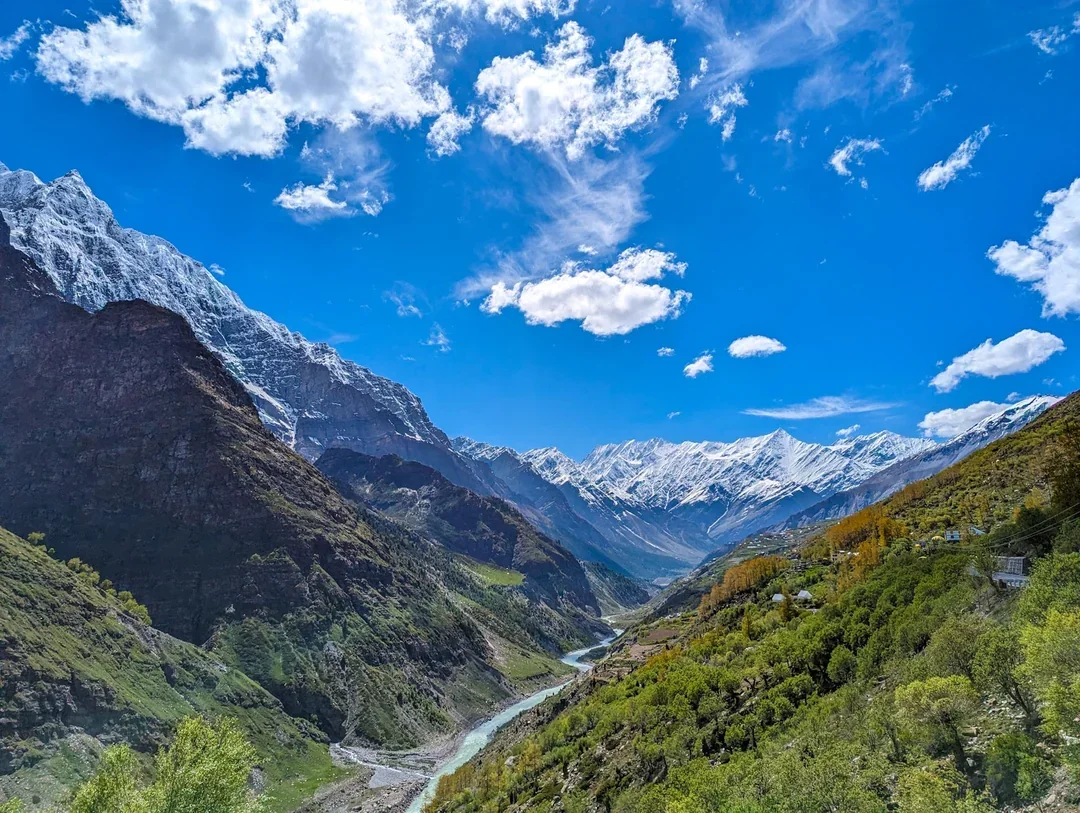 Snow-capped mountains under a blue sky with clouds, lush green valleys, and a river flowing through the valley.