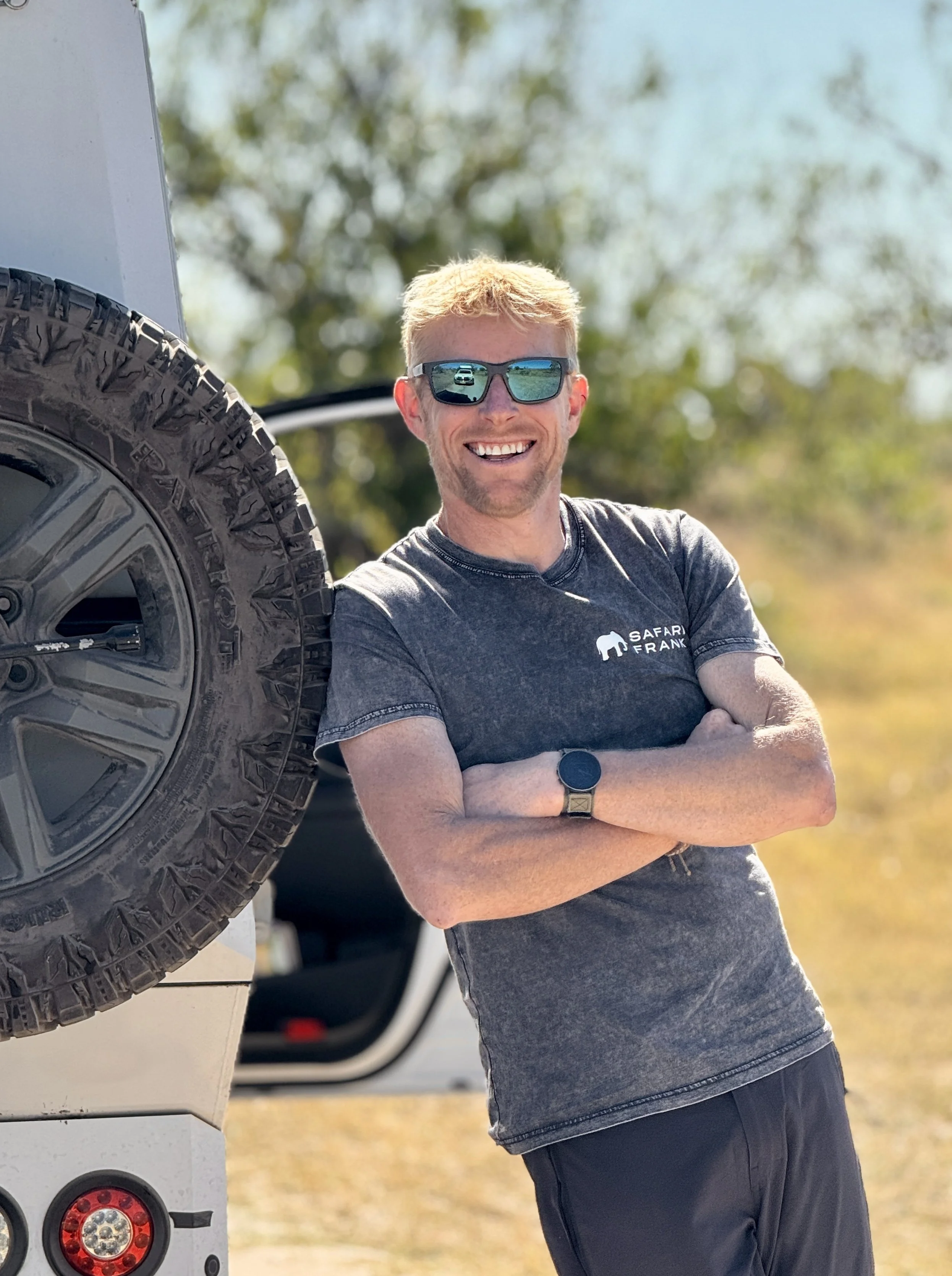 A smiling man with blond hair, wearing sunglasses and a dark gray T-shirt with a logo that says 'Safari Frank,' standing outdoors near a white vehicle with a large tire. He has crossed arms and is in a sunny environment with trees in the background.