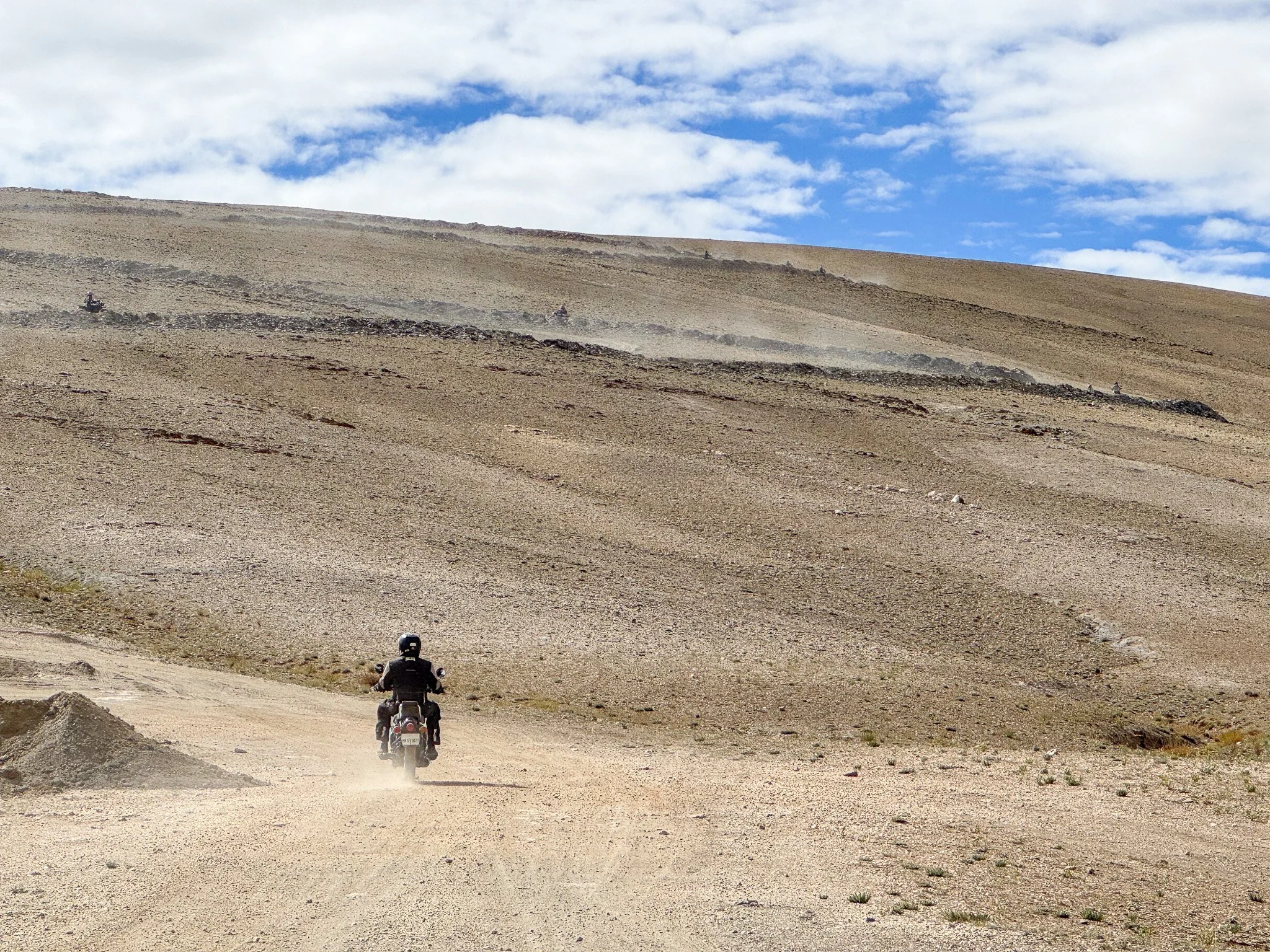 A person riding a motorcycle on a dirt trail in a barren, mountainous landscape under a partly cloudy sky.