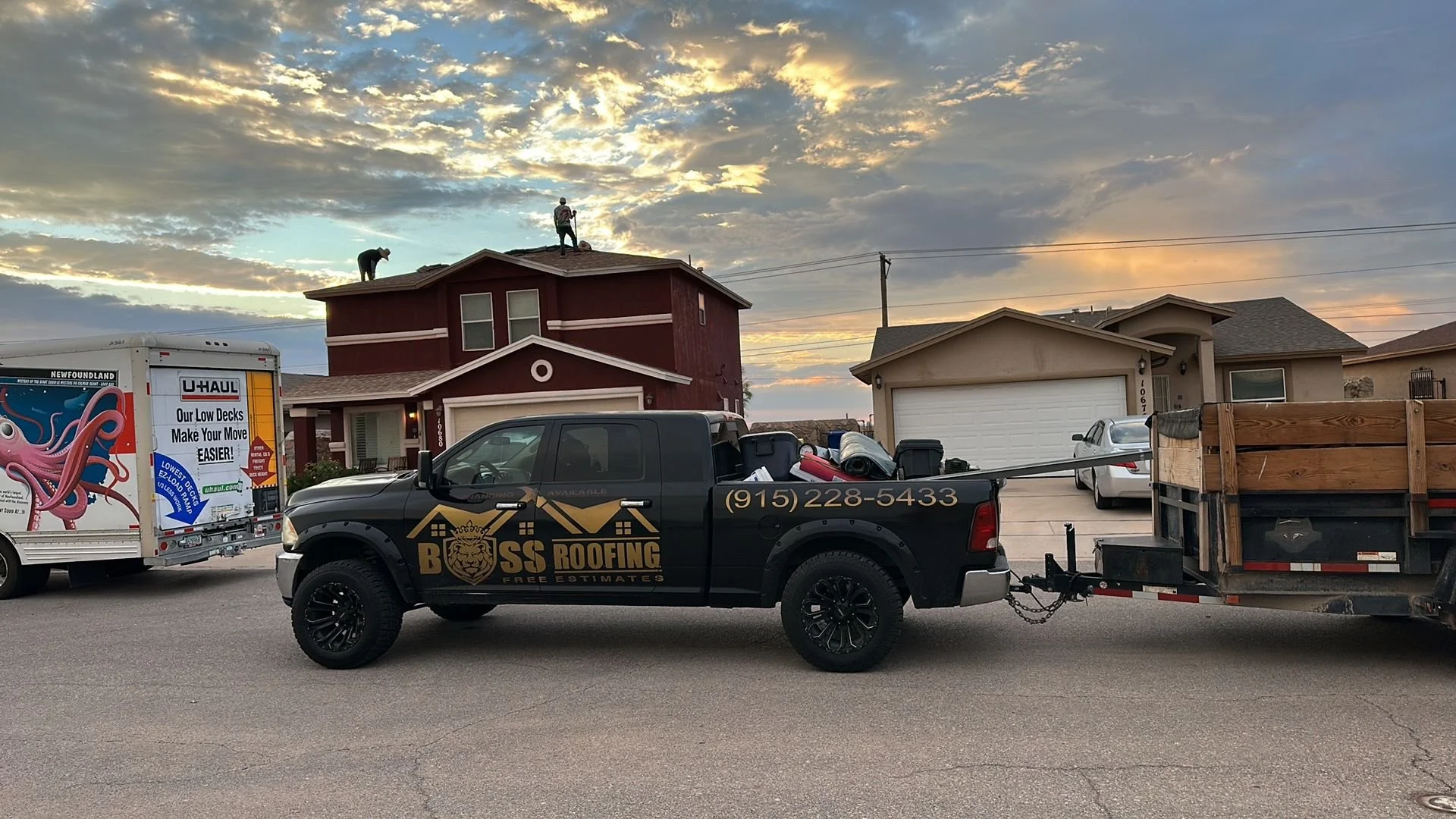 A black roofing company pickup truck with the logo and phone number, parked on a suburban street, with a house in the background and workers on the roof. There's a U-Haul truck nearby with an octopus advertisement, and the sky is cloudy at sunset.