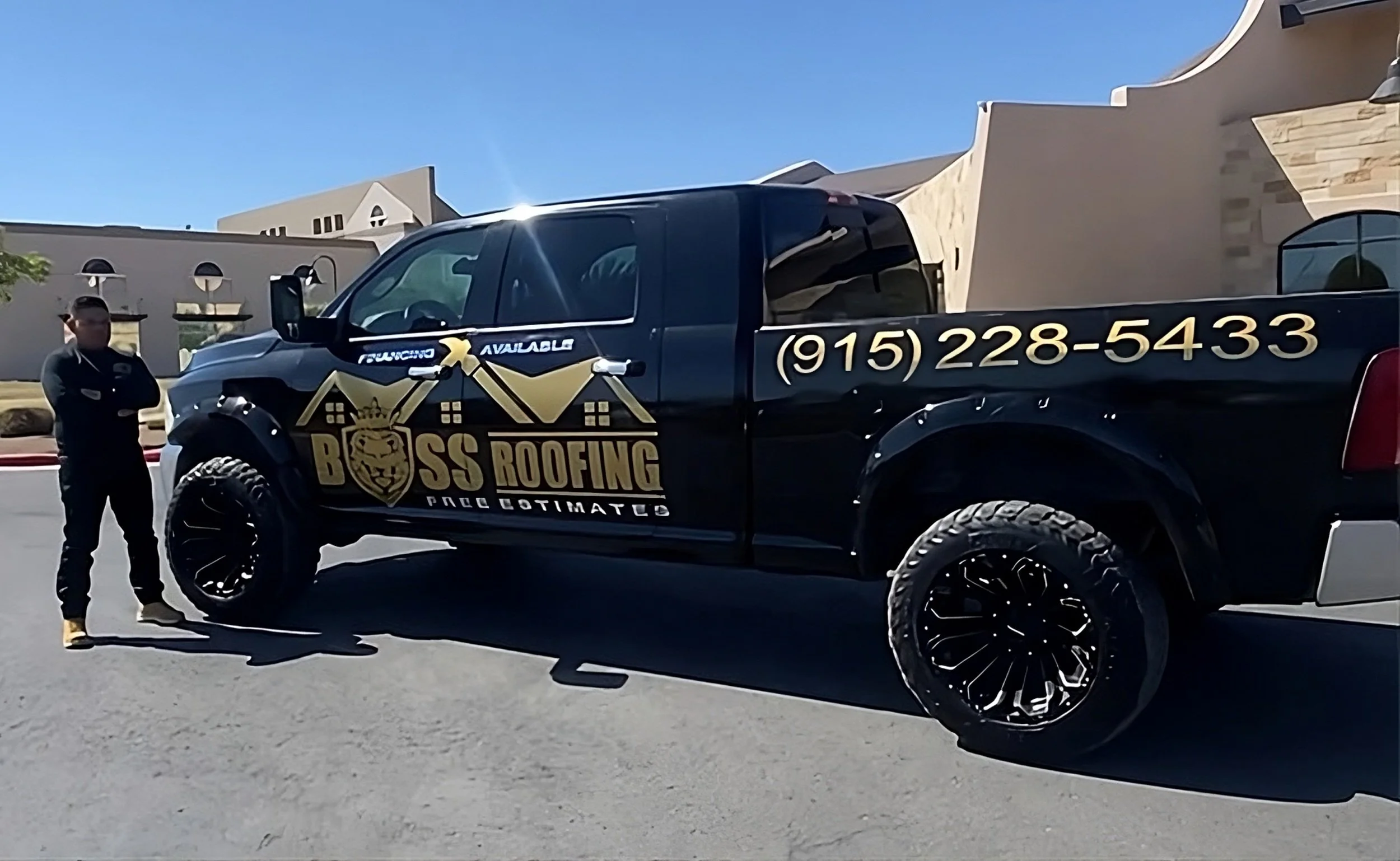 Black pickup truck with gold and white logo and text for Boss Roofing, and a man standing next to it with arms crossed. The truck has a phone number (915) 228-5433 and is parked on a street in front of buildings with a clear blue sky overhead.