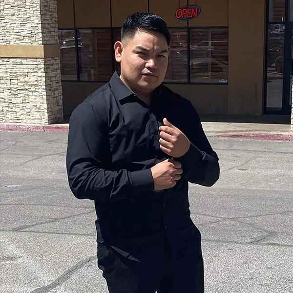 Man standing outside in front of a building with an 'OPEN' sign in the window, wearing a black shirt and making a fist with one hand.