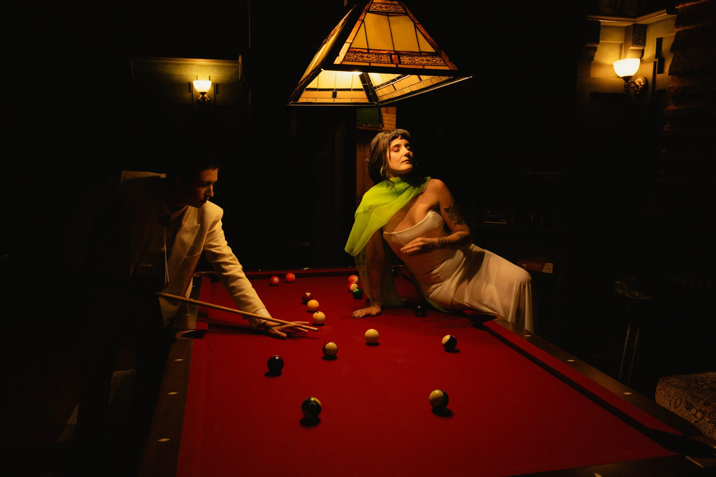 A woman in a white dress with a green scarf posing on a pool table at night, with a man in a beige suit preparing to take a shot, and warm lighting overhead.