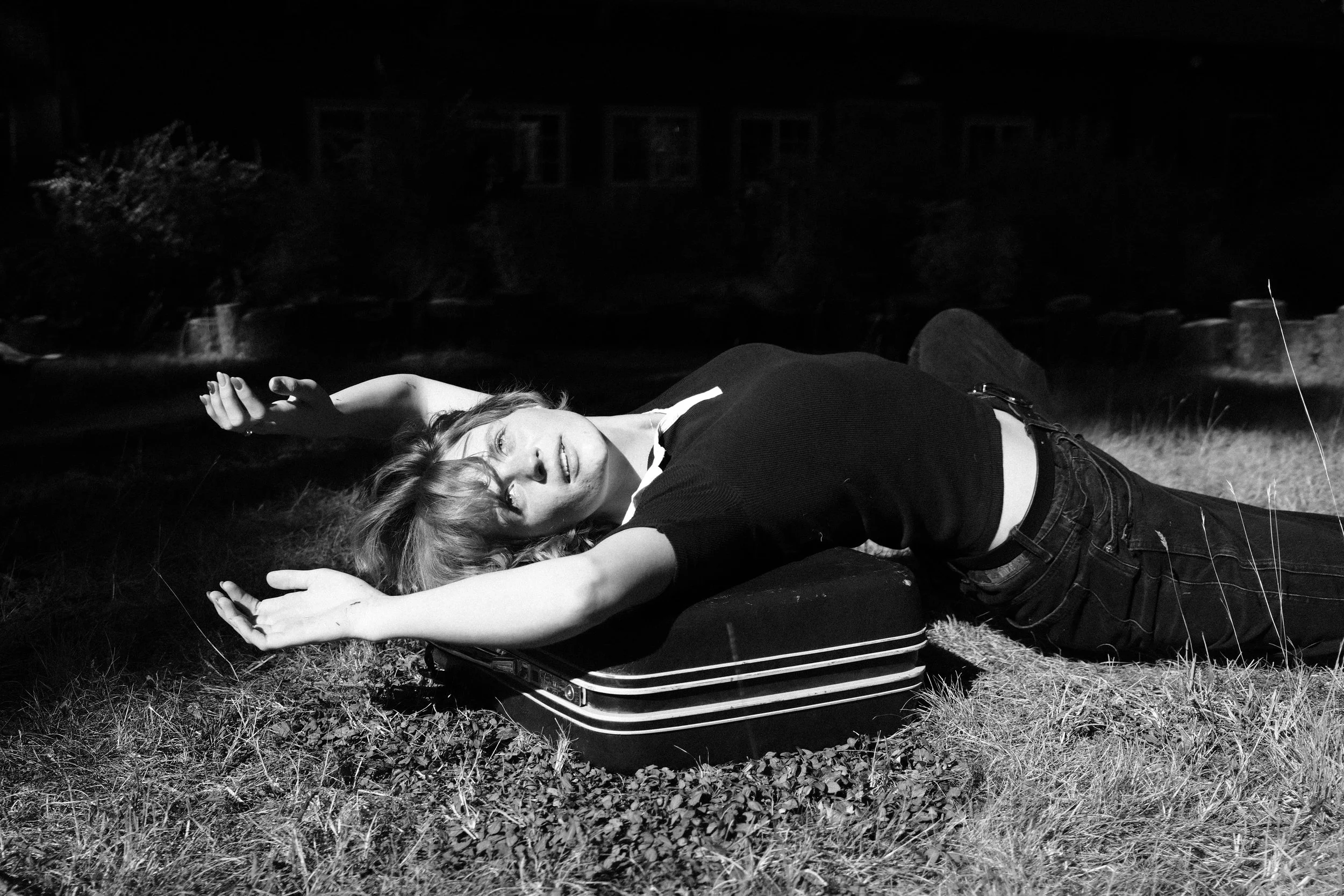 Creative black and white portrait of a woman lying on a suitcase in sunlight, photographed in a cinematic editorial style.