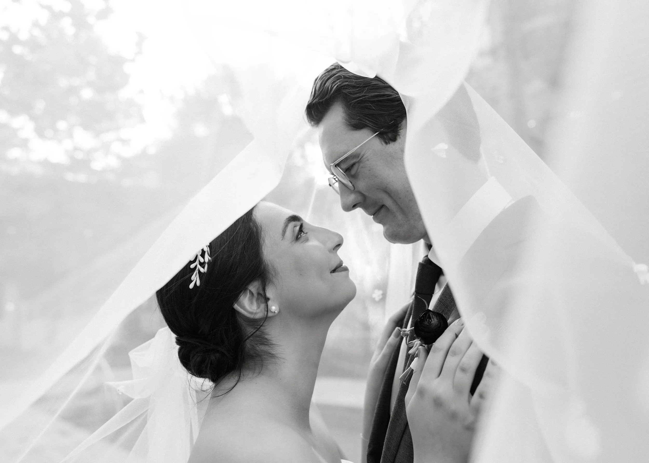 Black and white photo of a couple under a sheer veil, gazing at each other, with a blurred outdoor background.