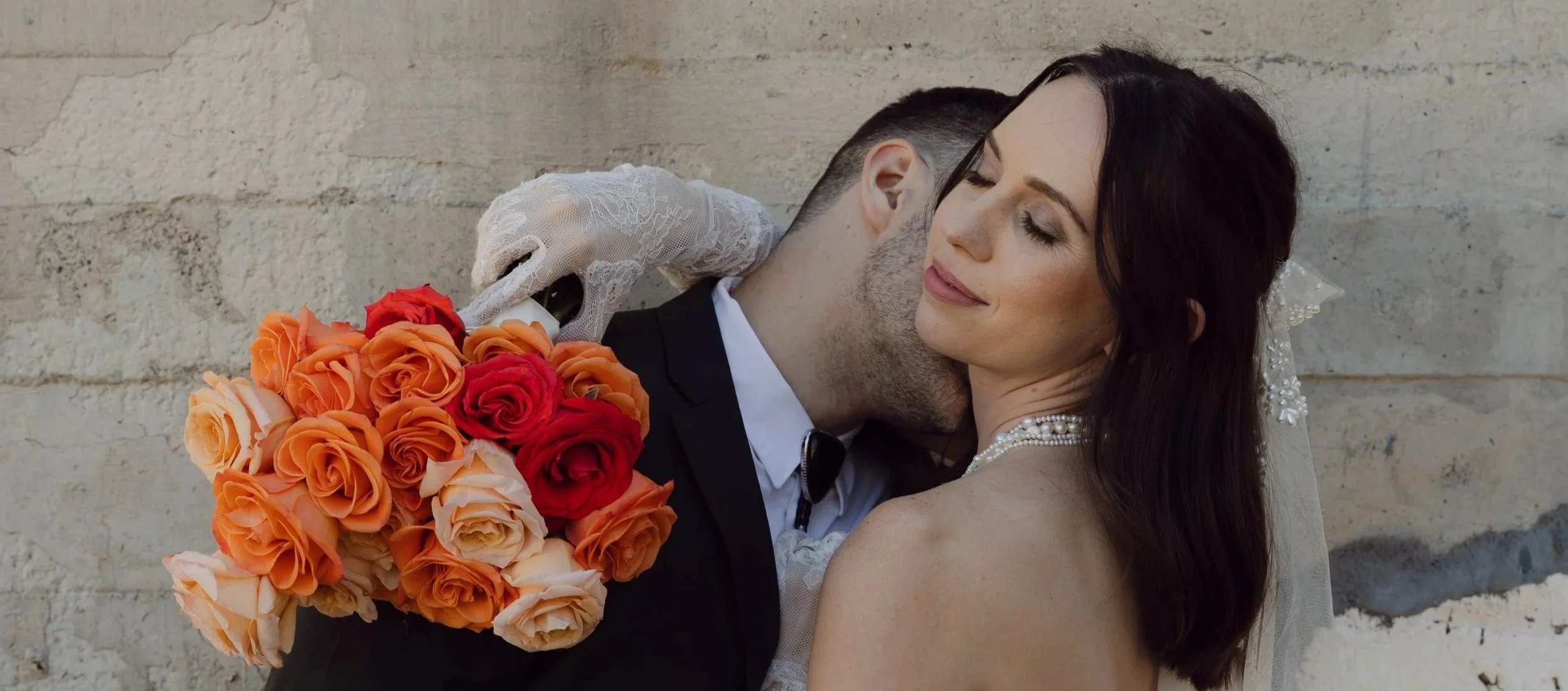 A bride and groom embrace, the groom kissing the bride's neck against a stone wall. She is smiling with her eyes closed, wearing a pearl necklace and veil. The groom is holding a bouquet of orange, red, and peach roses.