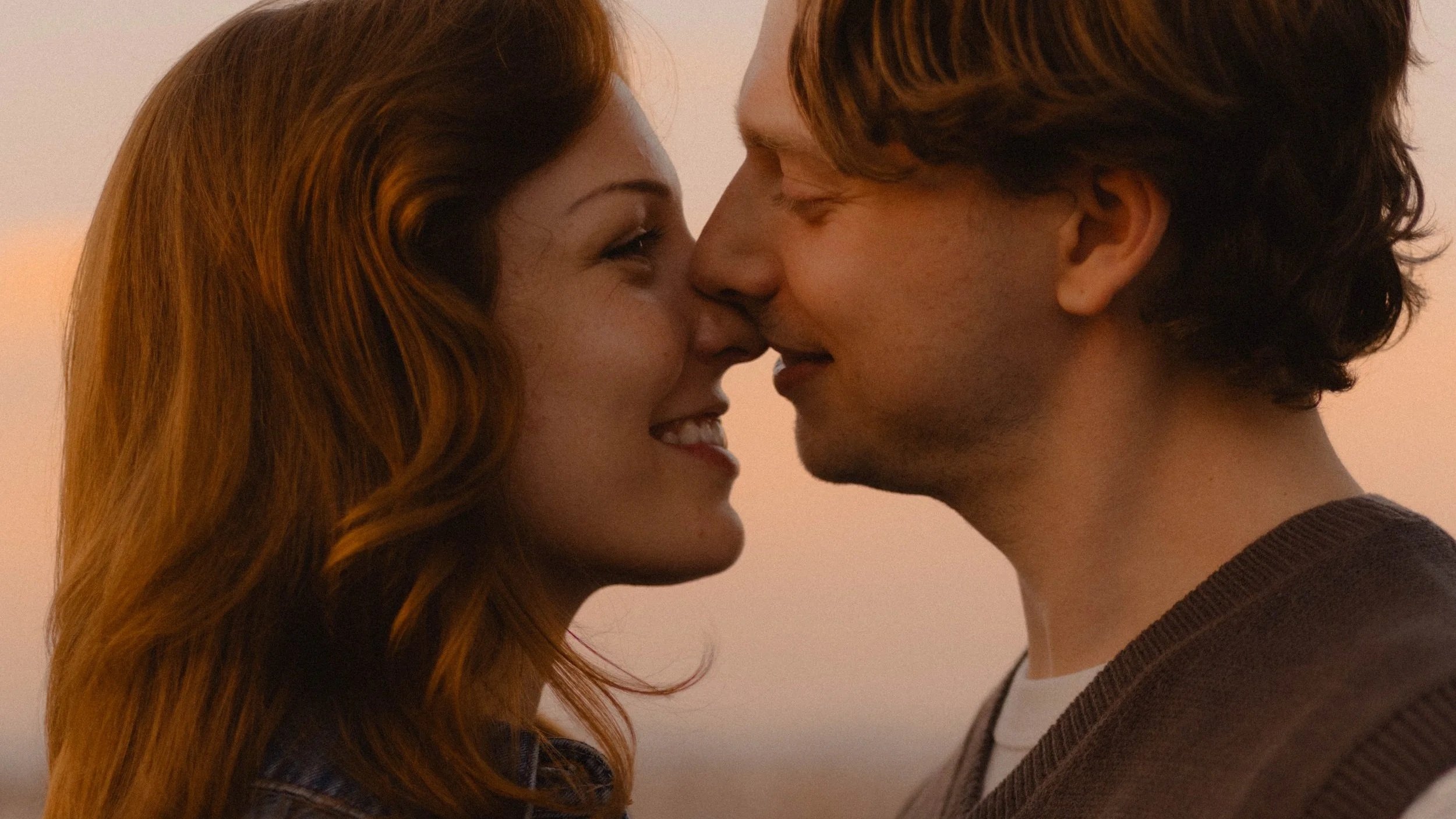 A man and woman close together, noses touching, smiling, in an intimate moment at sunset.
