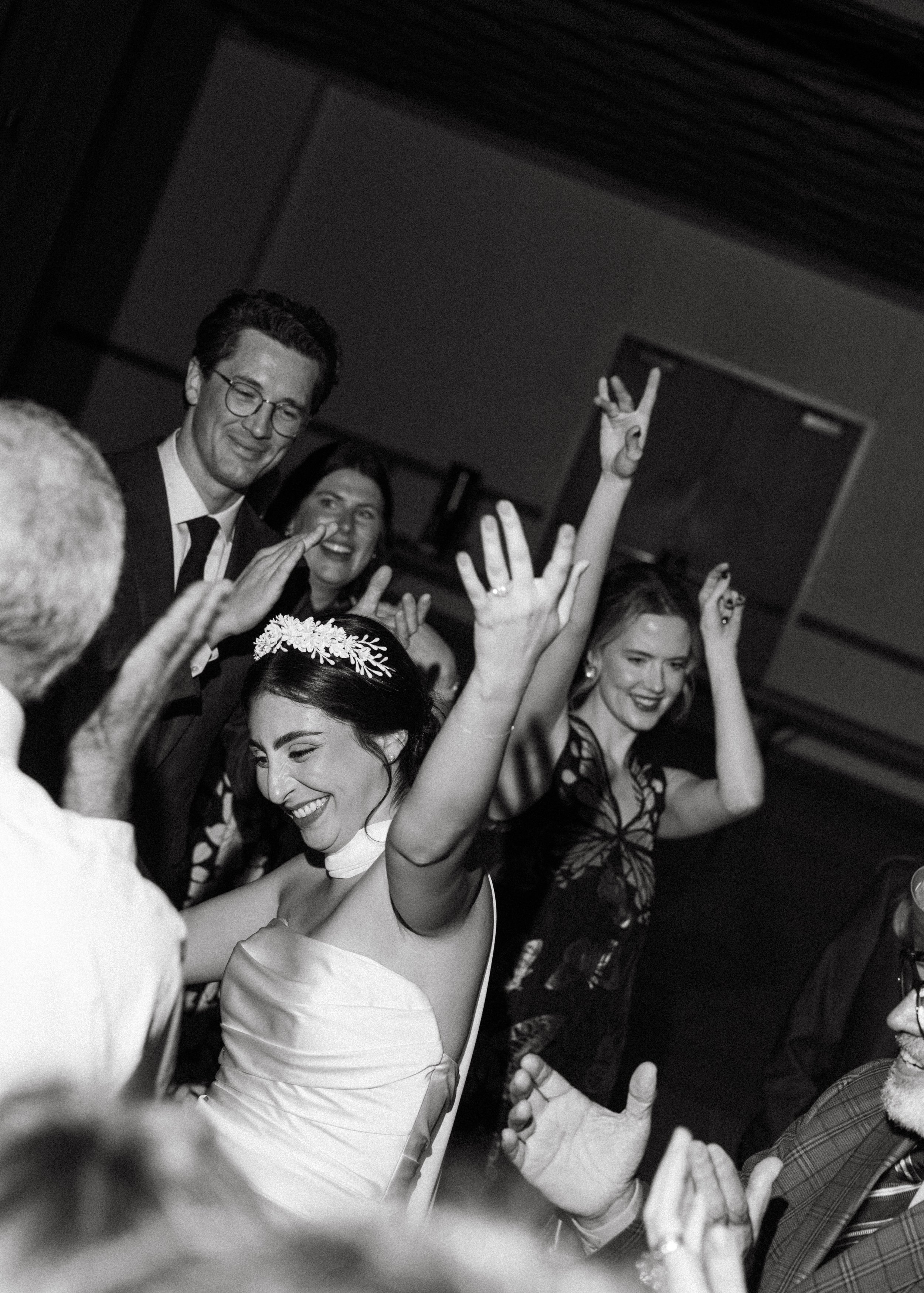 A woman in a wedding dress celebrating with friends and family at a wedding reception, smiling and raising her hands.