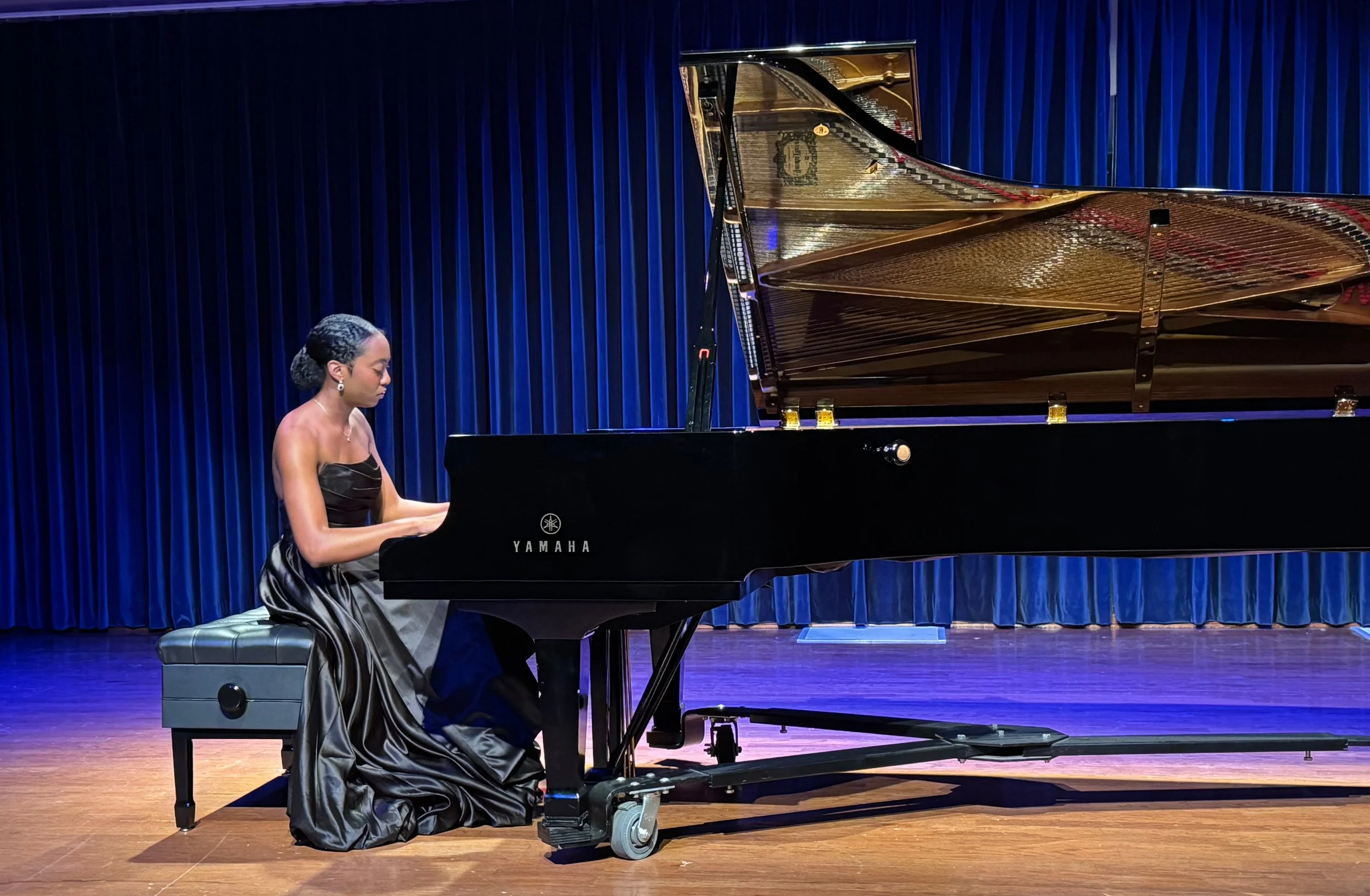 A woman in a black strapless dress playing a grand piano on a stage with blue curtains.