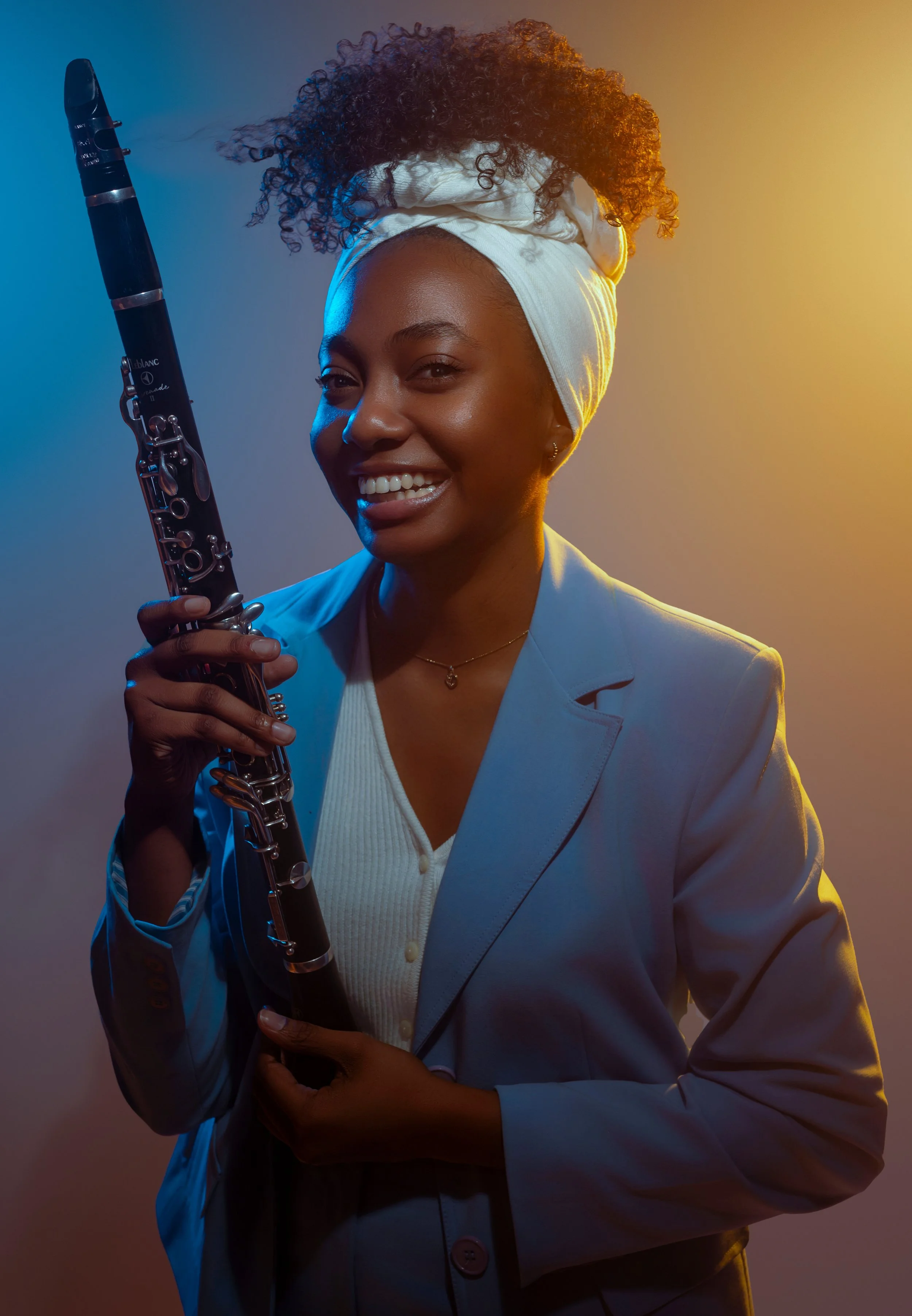 A woman wearing a white headwrap and blazer, holding a clarinet, smiling in front of a gradient background with blue and yellow lighting.