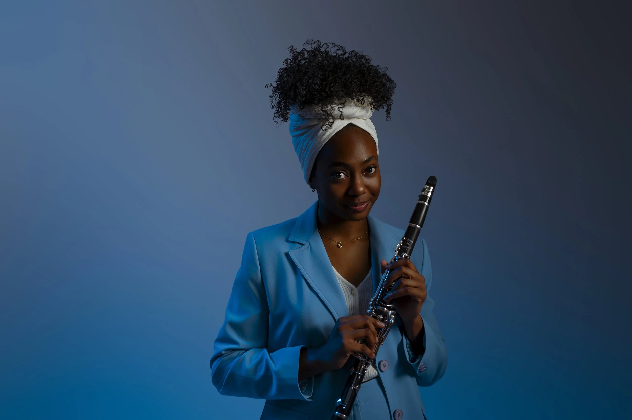 A woman with dark skin and curly hair wrapped in a white towel, wearing a blue blazer, holding a clarinet and smiling slightly, against a blue gradient background.