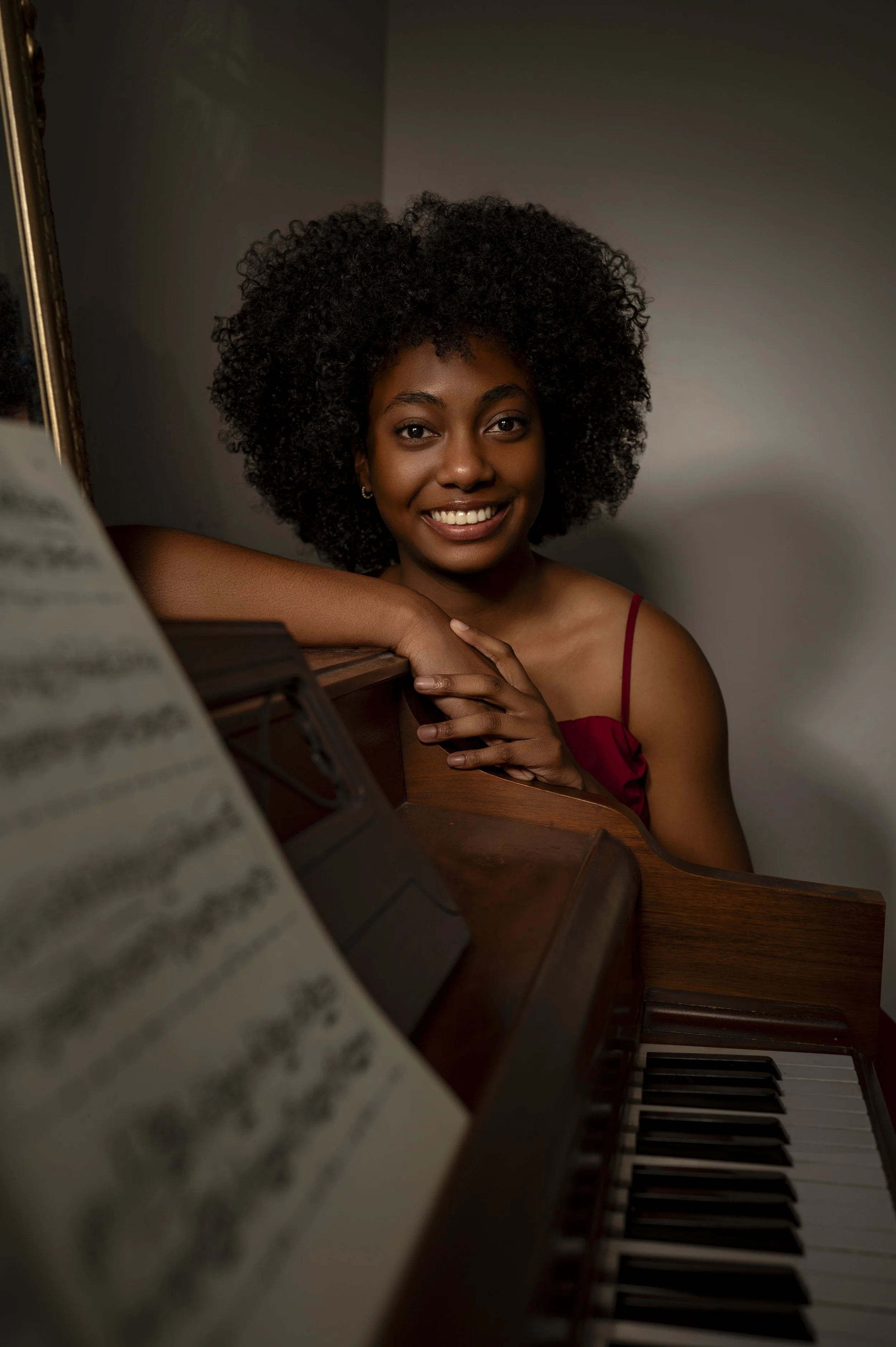 Young woman with curly black hair smiling, leaning on a piano with sheet music in the foreground.