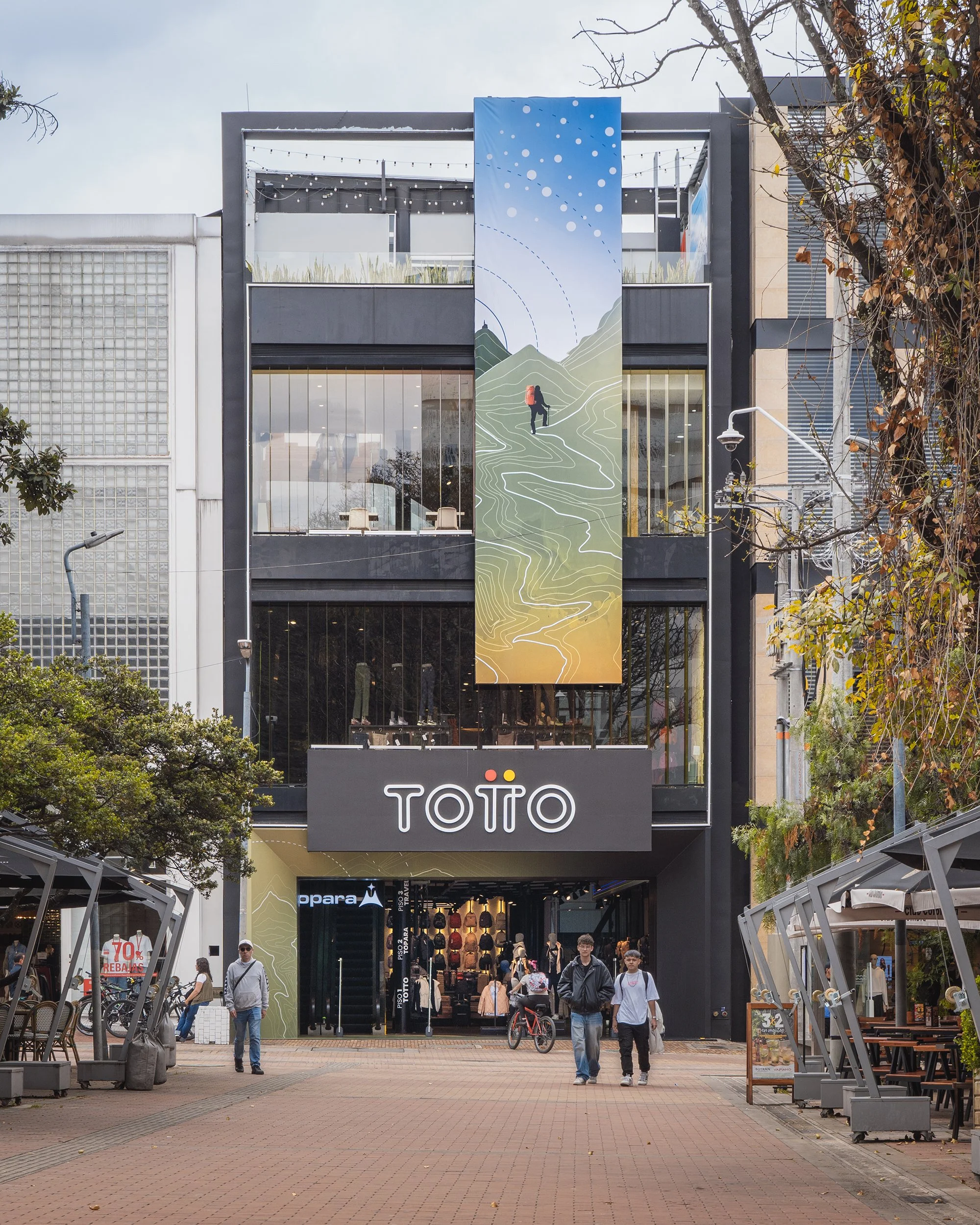 People walking out of a shopping mall on a busy day, with signage for Totto store on the building's facade and outdoor seating and bicycle racks in the foreground.