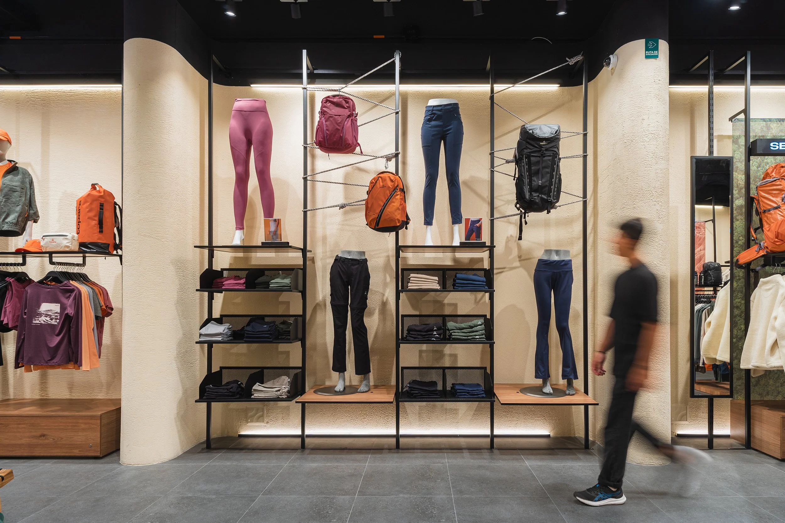 Display of outdoor clothing and gear in a retail store, including mannequins dressed in athletic pants, with backpacks and folded clothes on shelves behind.