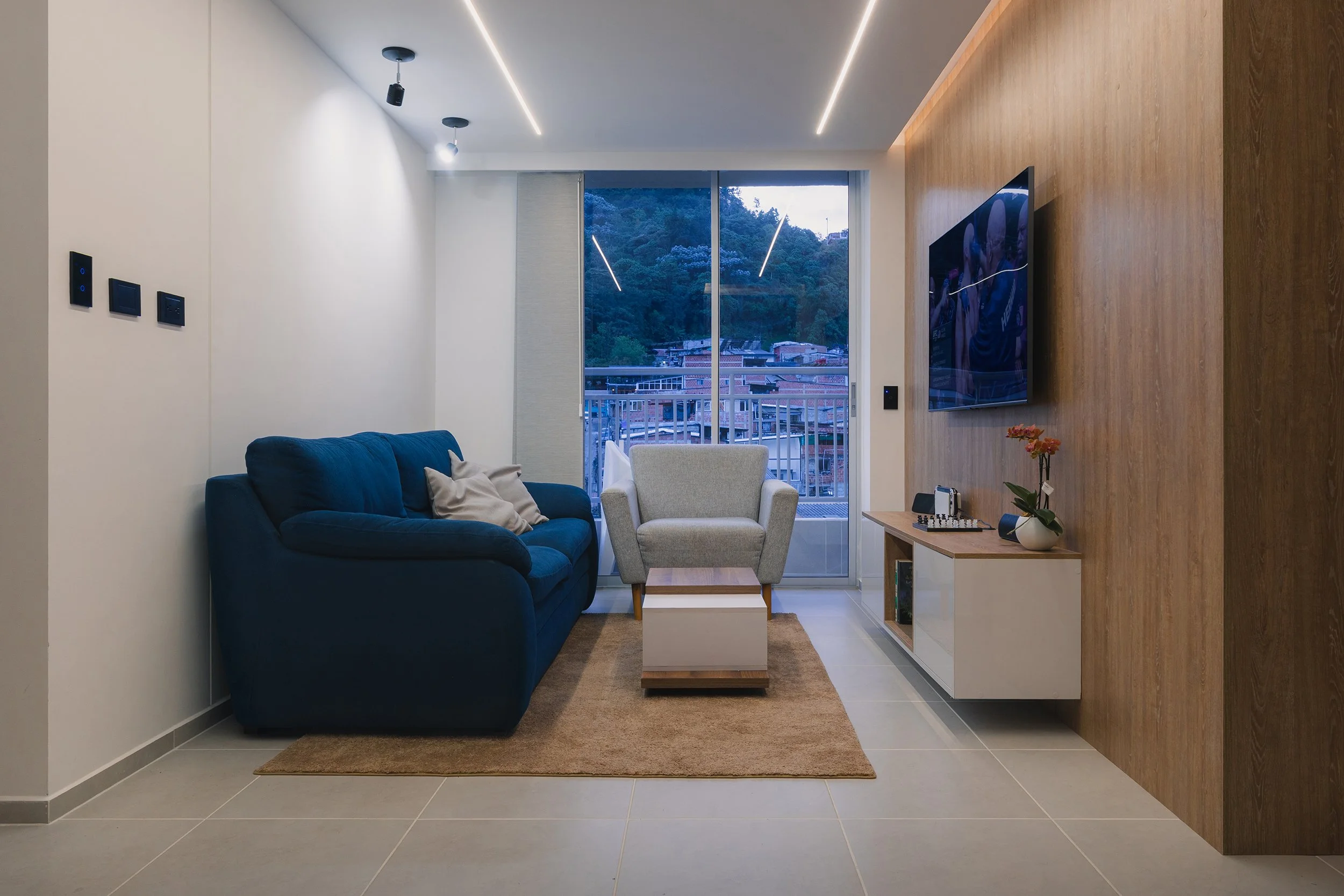 Modern living room with a navy blue sofa, a white armchair, a small coffee table, a flat-screen TV mounted on a wooden accent wall, and large sliding glass doors showing hills and buildings outside.