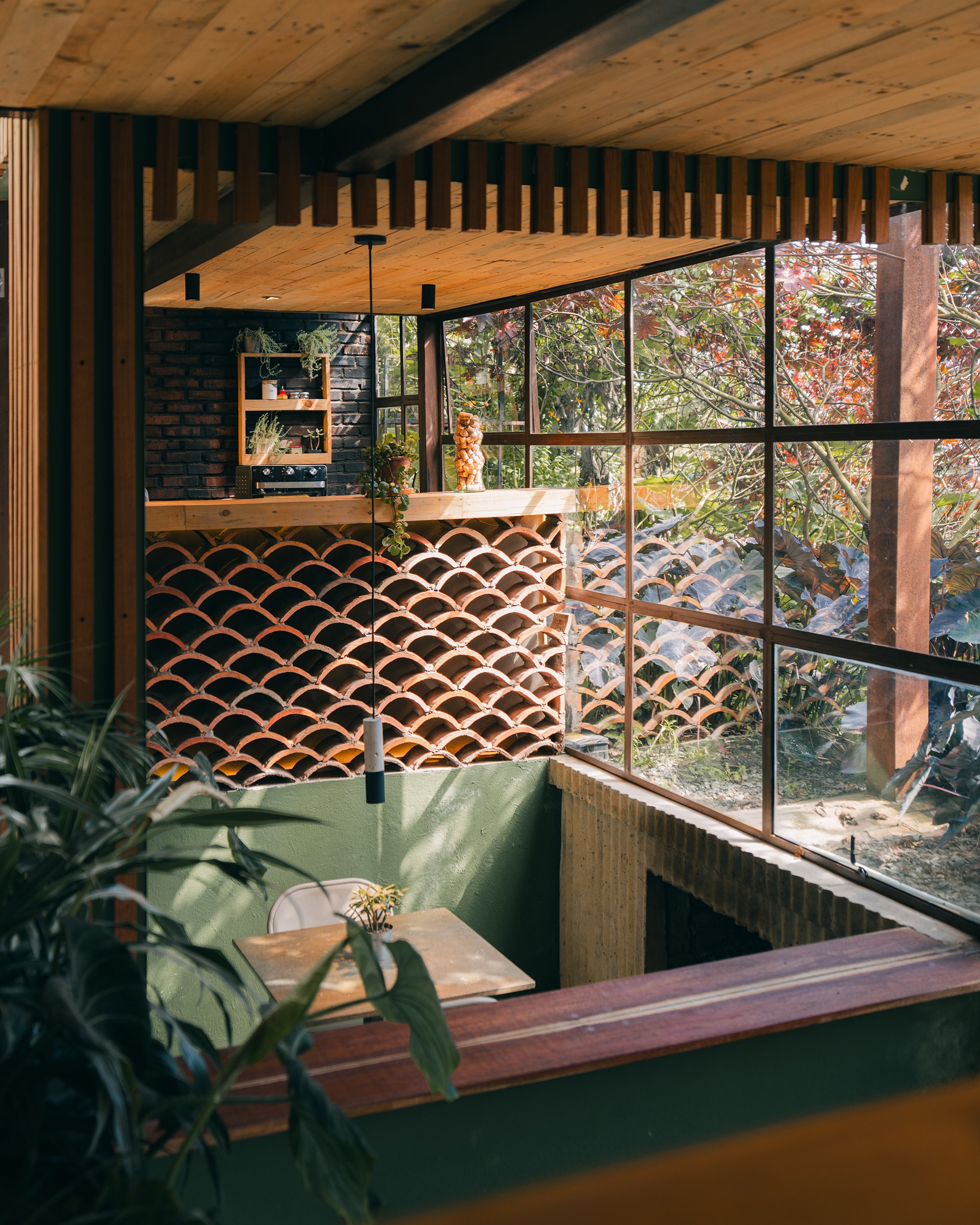Interior of a room with large windows showing trees outside, featuring wooden and brick decor, a small table with a plant, and a wine bottle display with firewood.