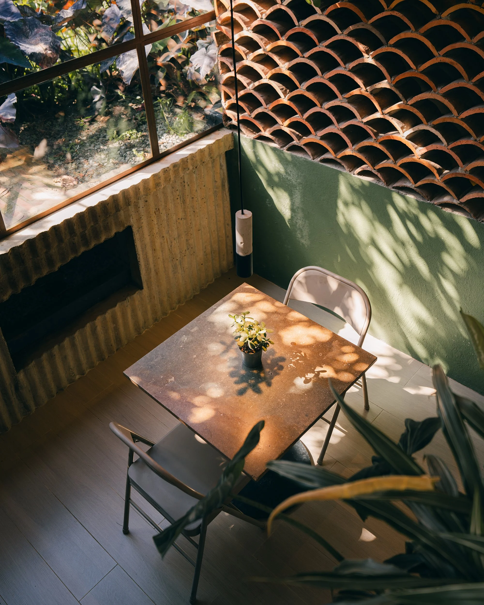 A cozy indoor space with a marble dining table and metal chairs, illuminated by sunlight and shadows, next to a window with a view of a garden with plants and leaves outside.