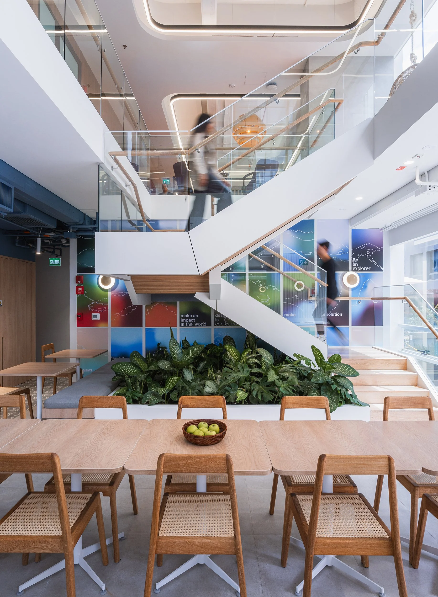 Modern multi-level office or lounge area with stairs, glass railings, green plants, and colorful wall art.