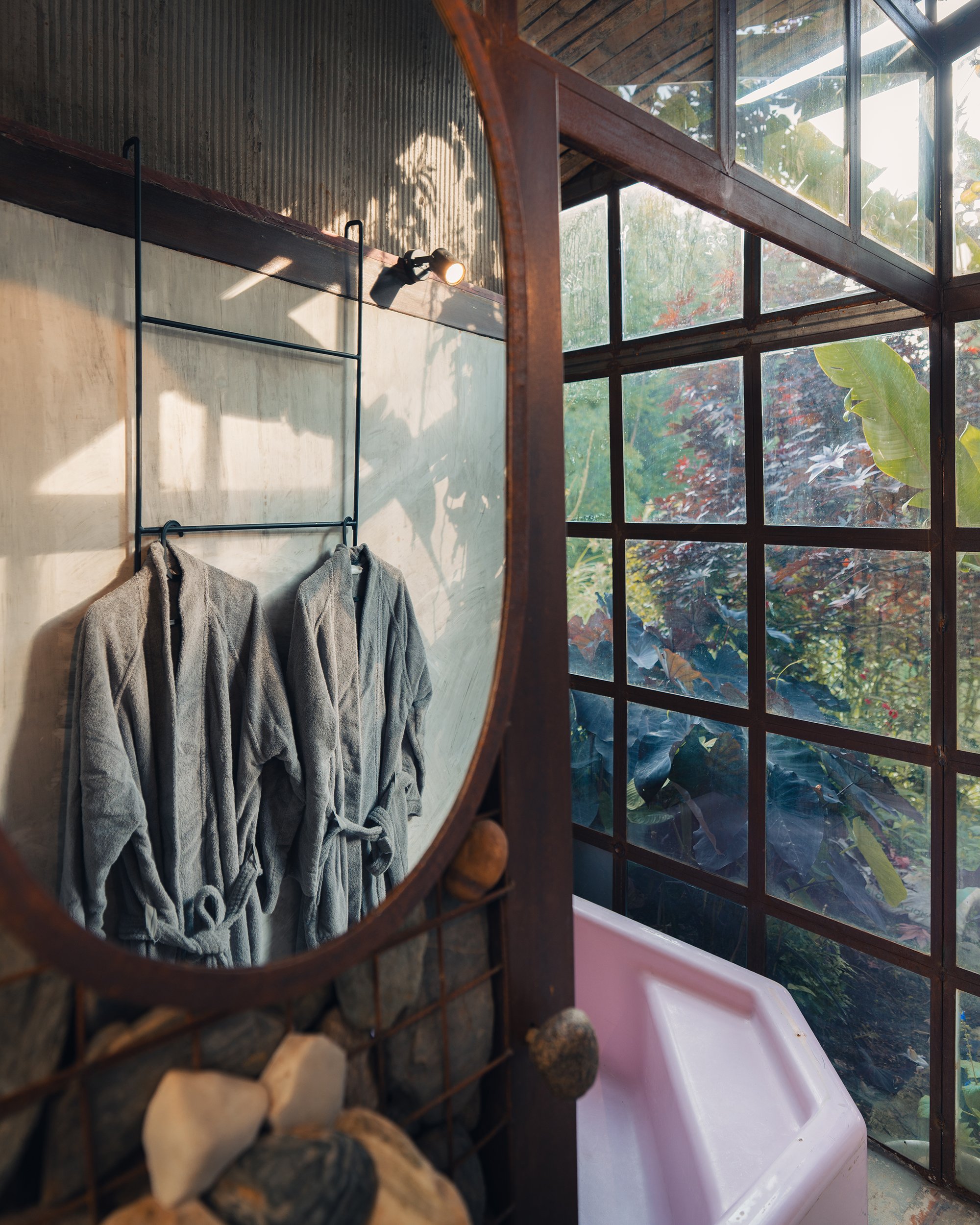 Interior view of a house with large glass windows showing green foliage outside. The interior features a rustic wall with a spotlight and hanging towels, and a pile of firewood stacked beside a mirror.