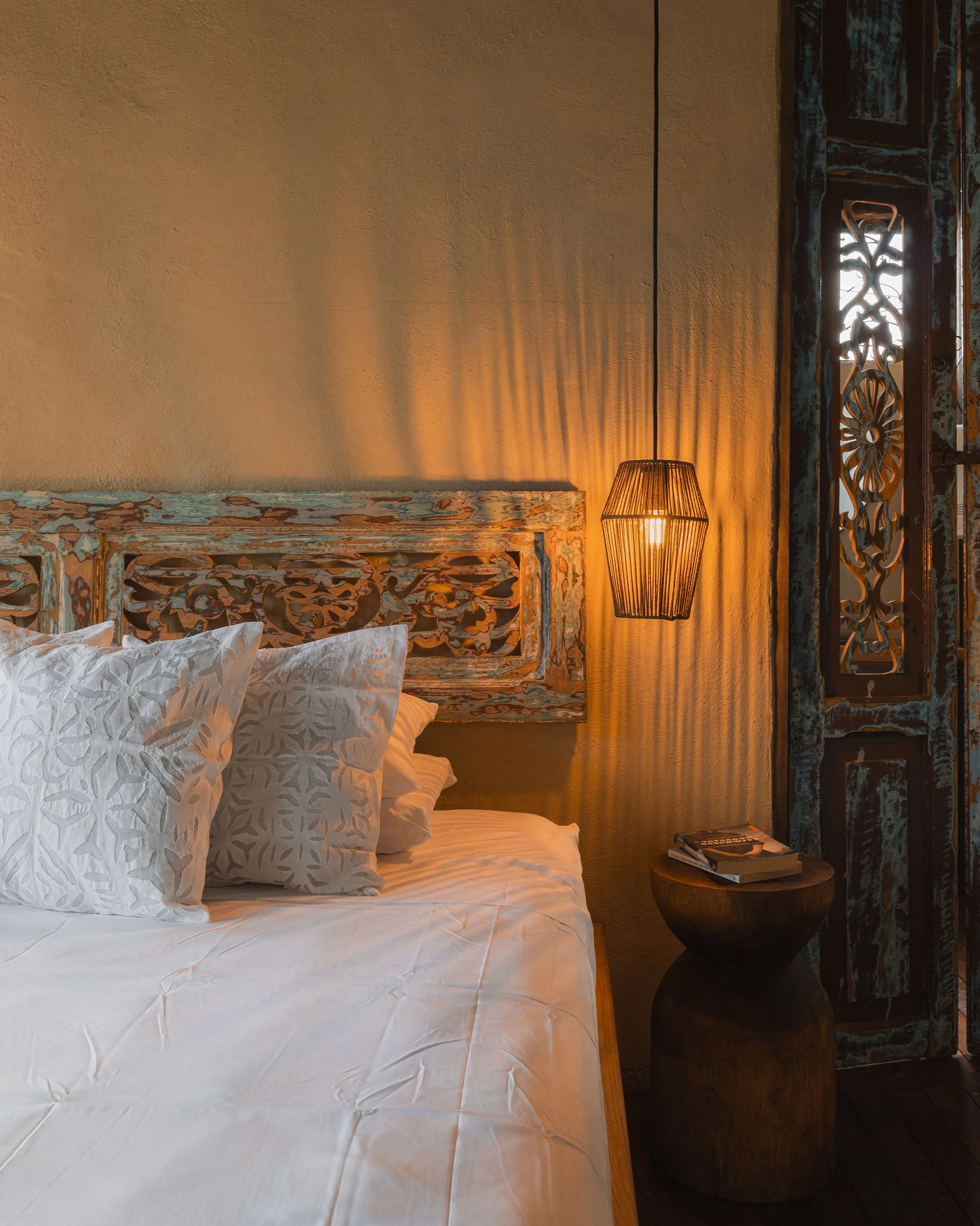 Fotografía de interiores del detalle del espaldar de una cama, que se encuentra hecho en madera tallada con formas organizas. Lampara colgante y mesa de noche