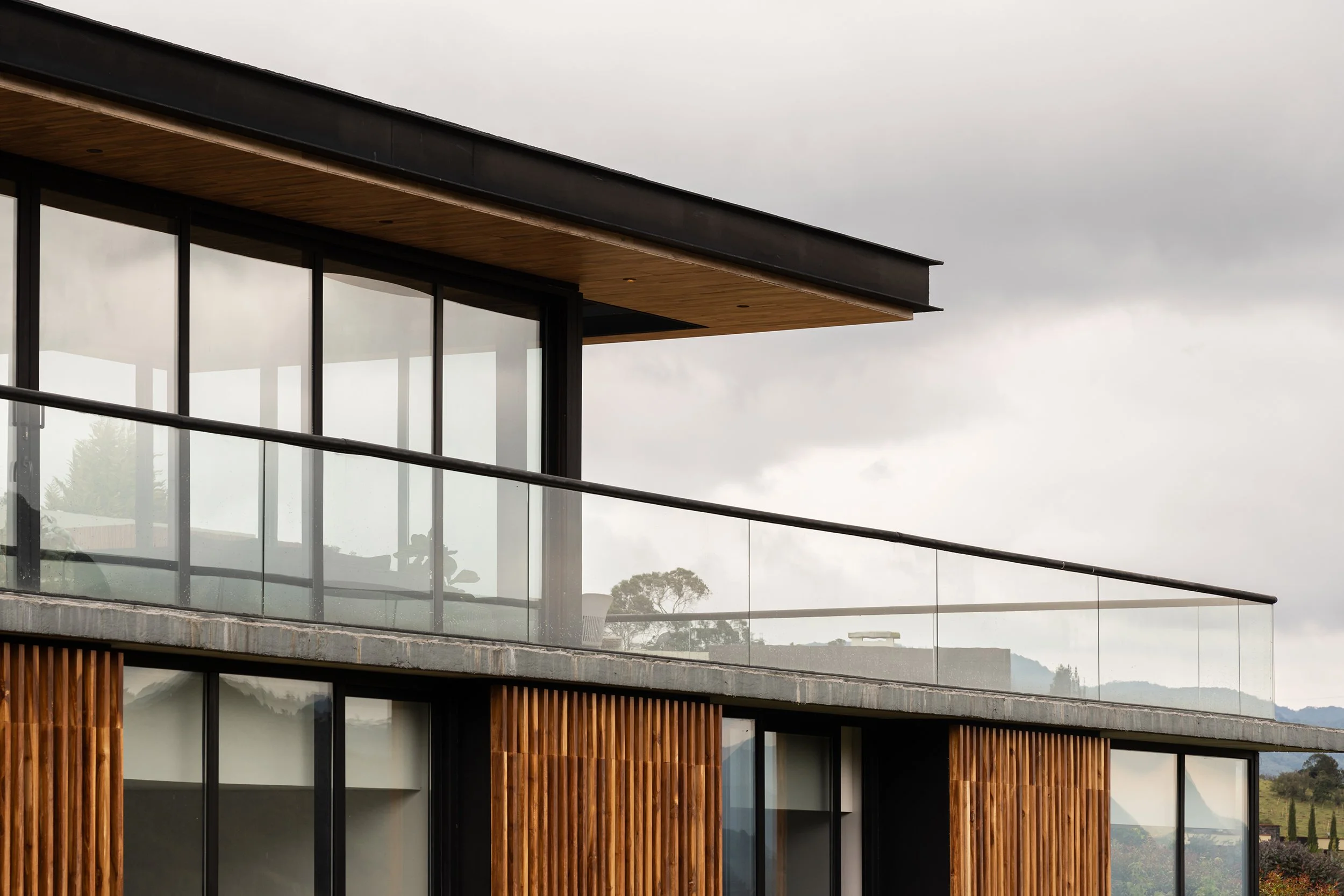 Fotografía profesional de arquitectura de una vivienda de dos niveles, con acabados en concreto a la vista, vigas metálicas con pintura negra, listones de madera y una terraza semicubierta con pasamanos en vidrio